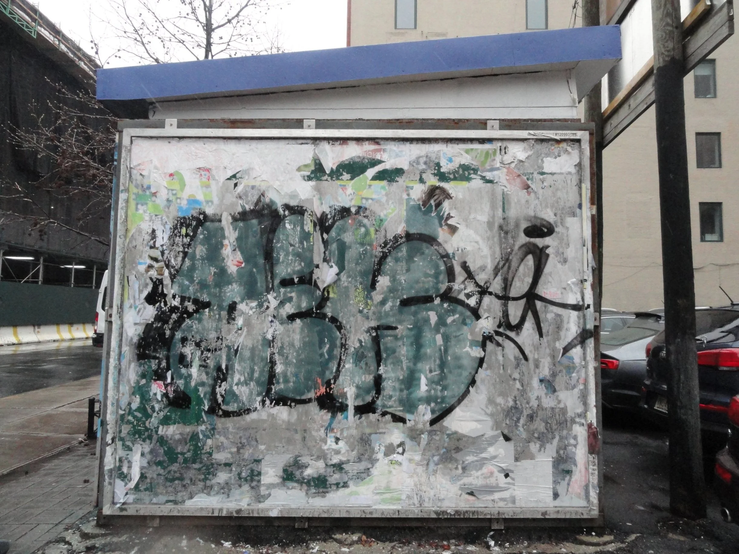 Weathered and torn advertising or poster board covered with graffiti and partially peeled stickers, situated on a city sidewalk, with parked cars and buildings in the background.