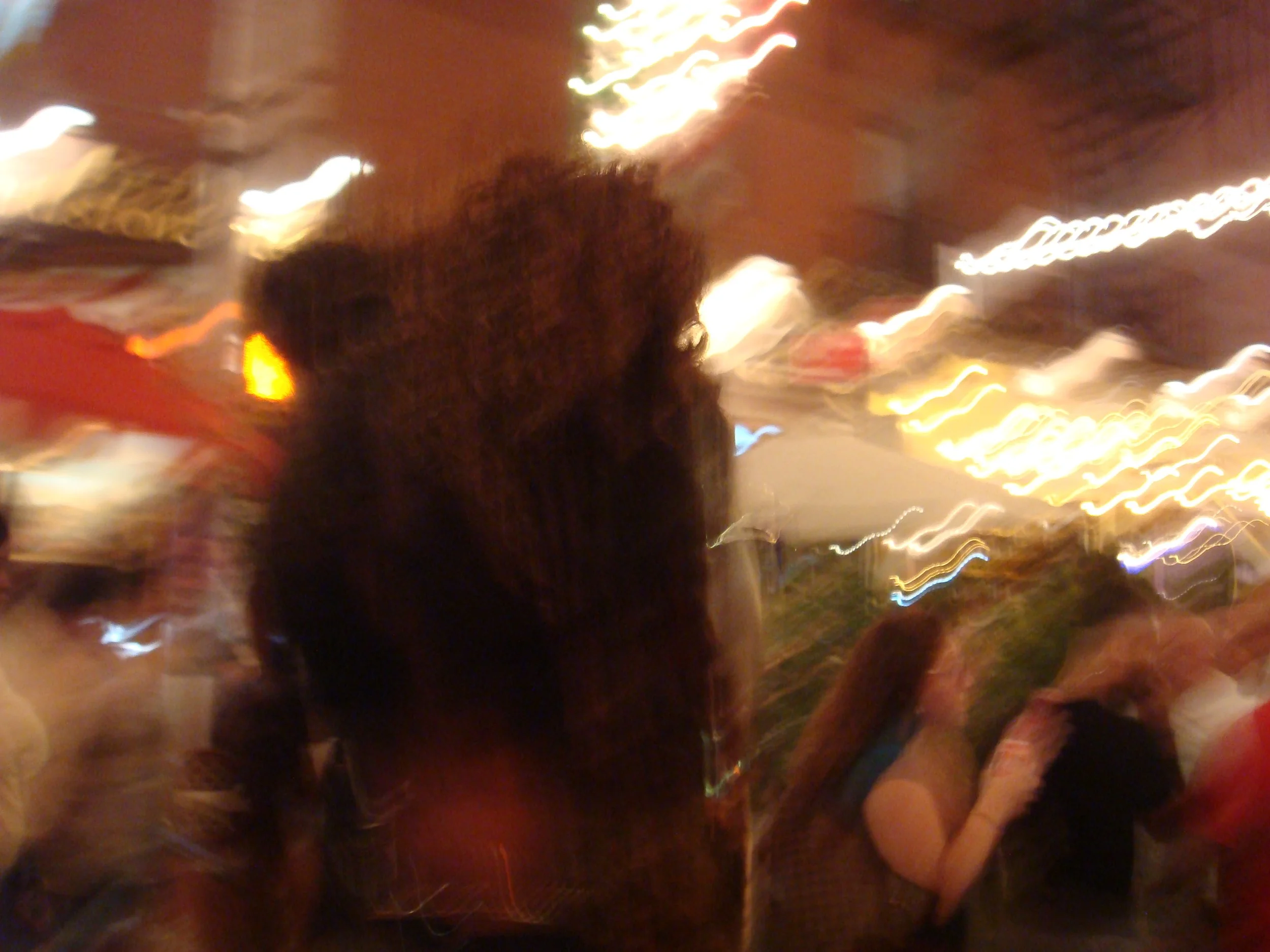 A blurry photo of a person with long hair in a crowded outdoor area at night, with colorful blurred lights and people in the background.