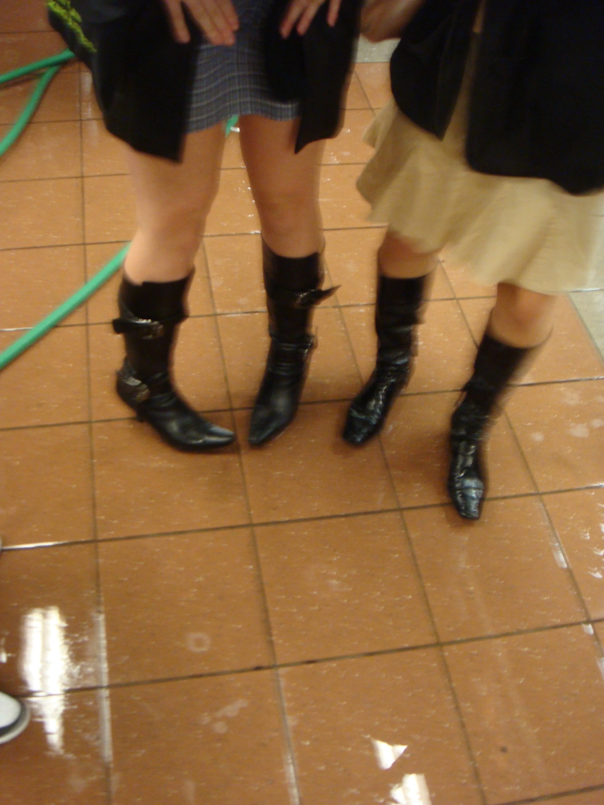 Two women wearing black knee-high boots, standing on a wet tiled floor, with one wearing a plaid skirt and the other a beige skirt.