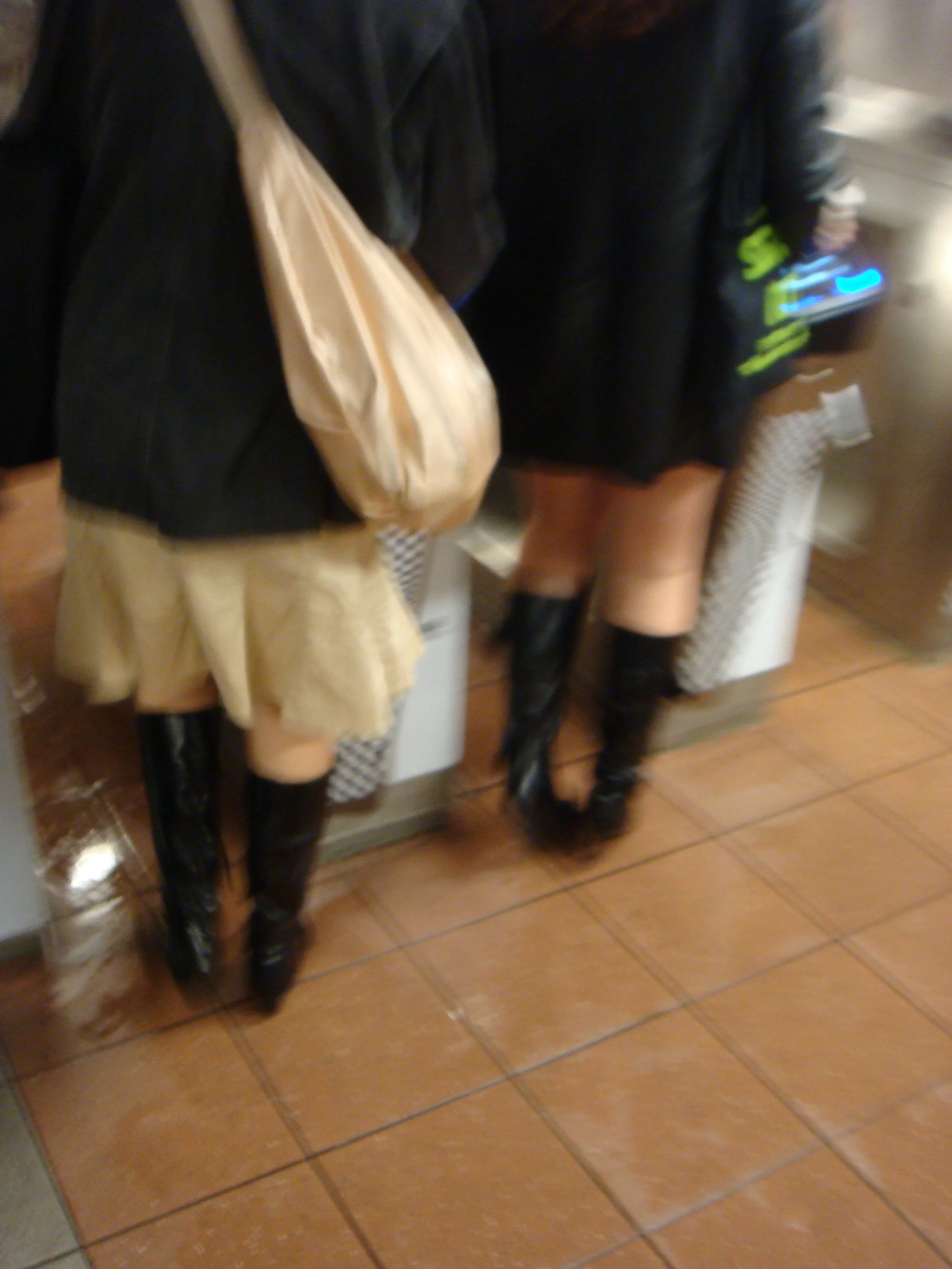 Two women in black shorts and over-the-knee black boots, standing at a counter. One is wearing a black jacket and a beige skirt; the other is wearing a black jacket with neon green writing and is holding a blue-lit smartphone.