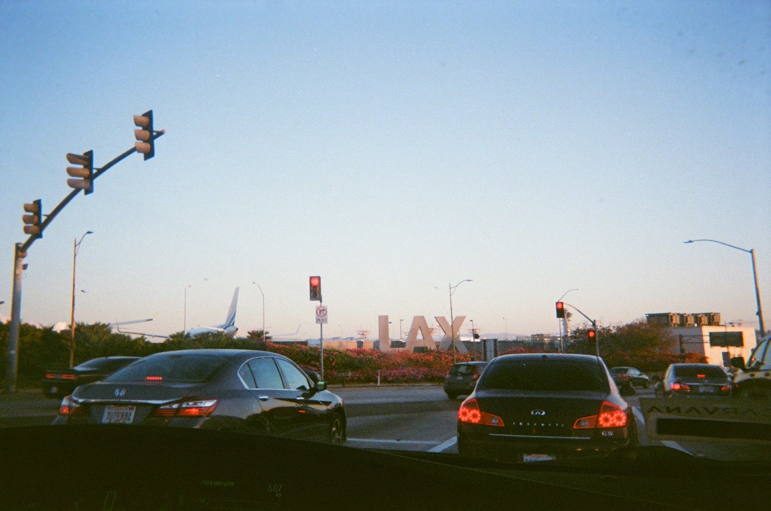 Traffic light with red signals, cars on the road, and large letters spelling 'LAX' in the background indicating Los Angeles Airport at sunset.