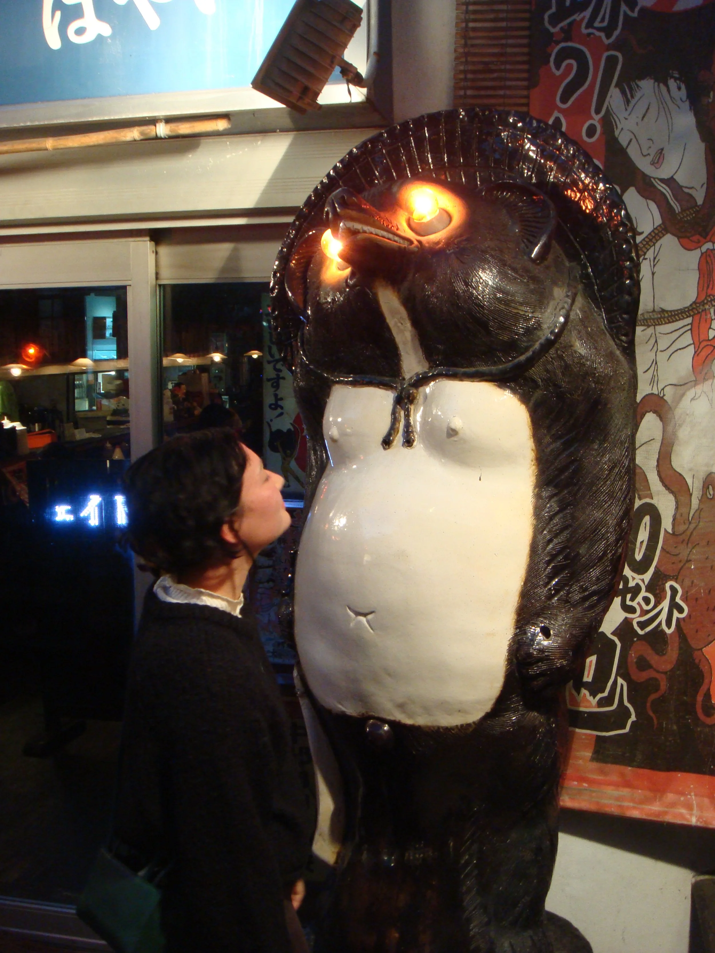 A woman standing in front of a large black and white bear sculpture with candles on its head, inside a building with Japanese signs and decor.