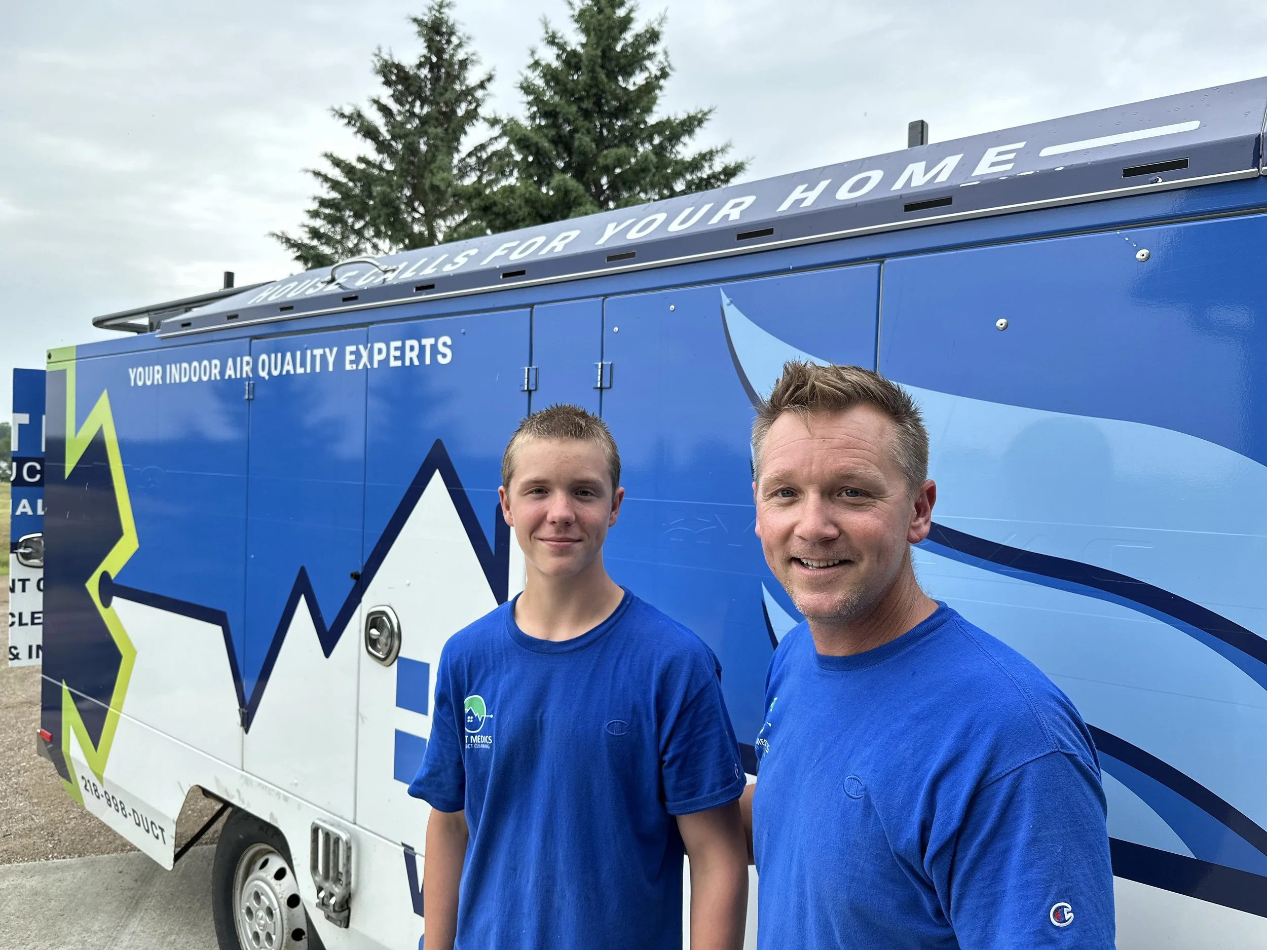 Two smiling men, one younger and one older, standing in front of a blue mobile air quality testing van with marketing graphics and text. Both are wearing matching blue T-shirts.