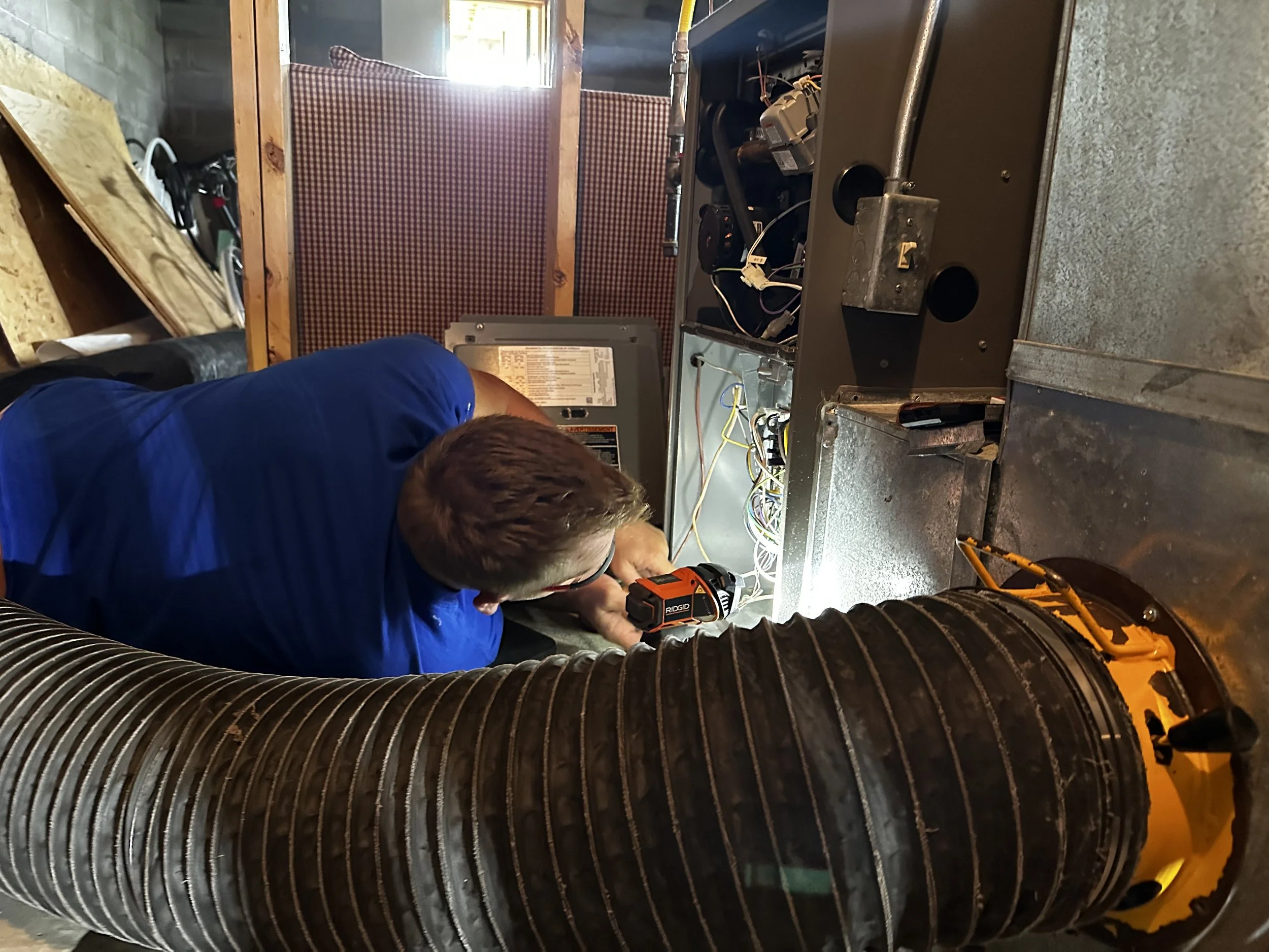 A man working inside a furnace or HVAC unit, using a cordless drill to adjust or repair the system, with various wires and components visible around him.