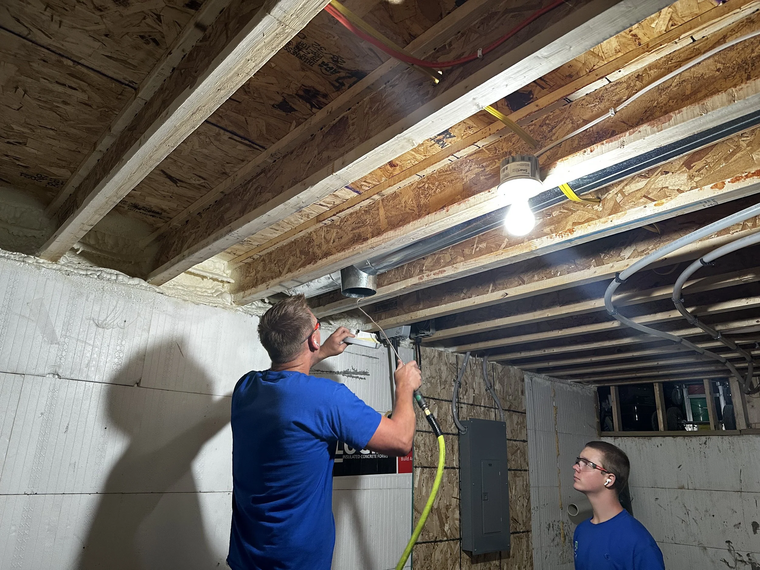 Two men working on electrical wiring in a construction site, with unfinished wood and drywall around, and a bright light bulb hanging from the ceiling.