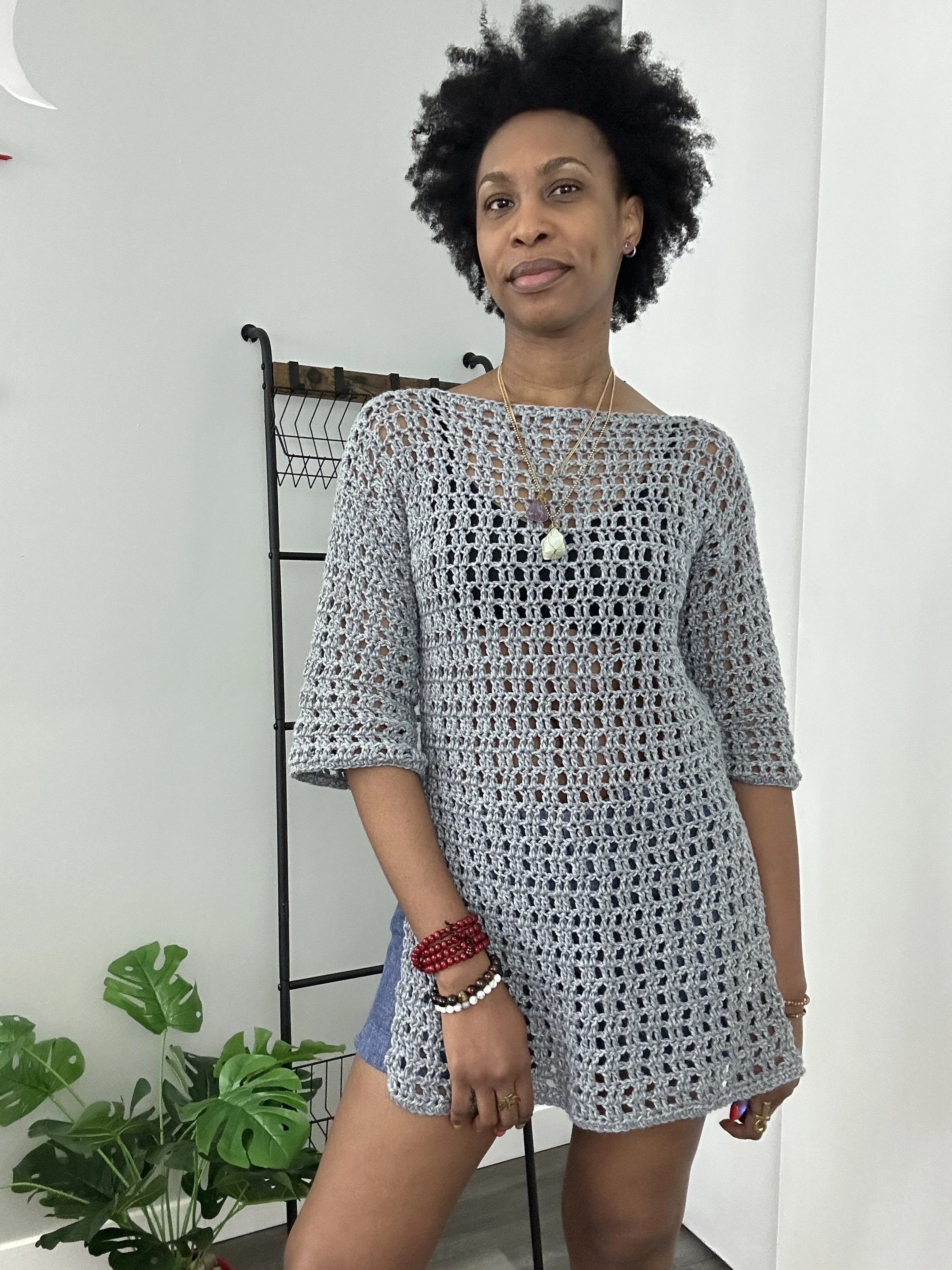 A woman with natural curly hair wearing a crochet top, multiple necklaces, and beaded bracelets, standing in front of a white wall with some greenery and a black metal rack in the background.