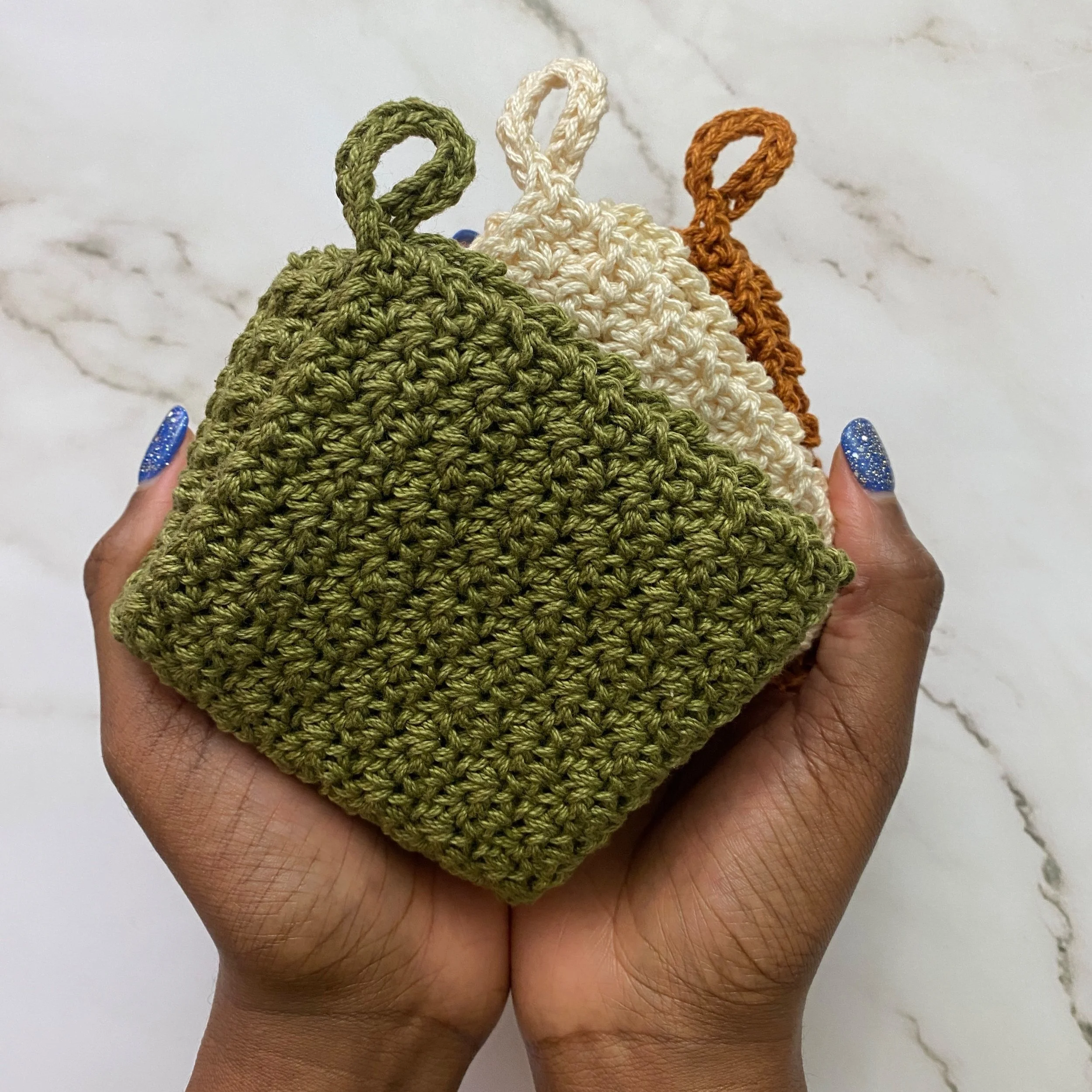 Three folded crochet washcloths in green, white, and orange, held between fingers with blue glittery nails, against a white marble background.
