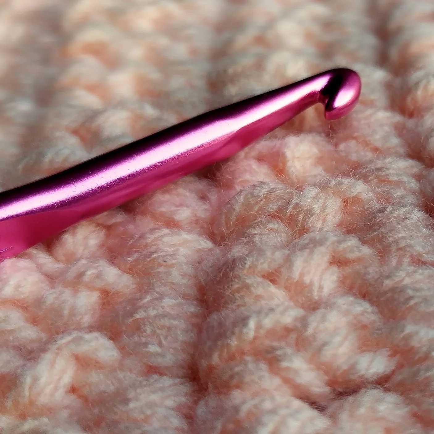 Close-up of a pink crochet hook resting on pink and beige chunky knitted fabric.