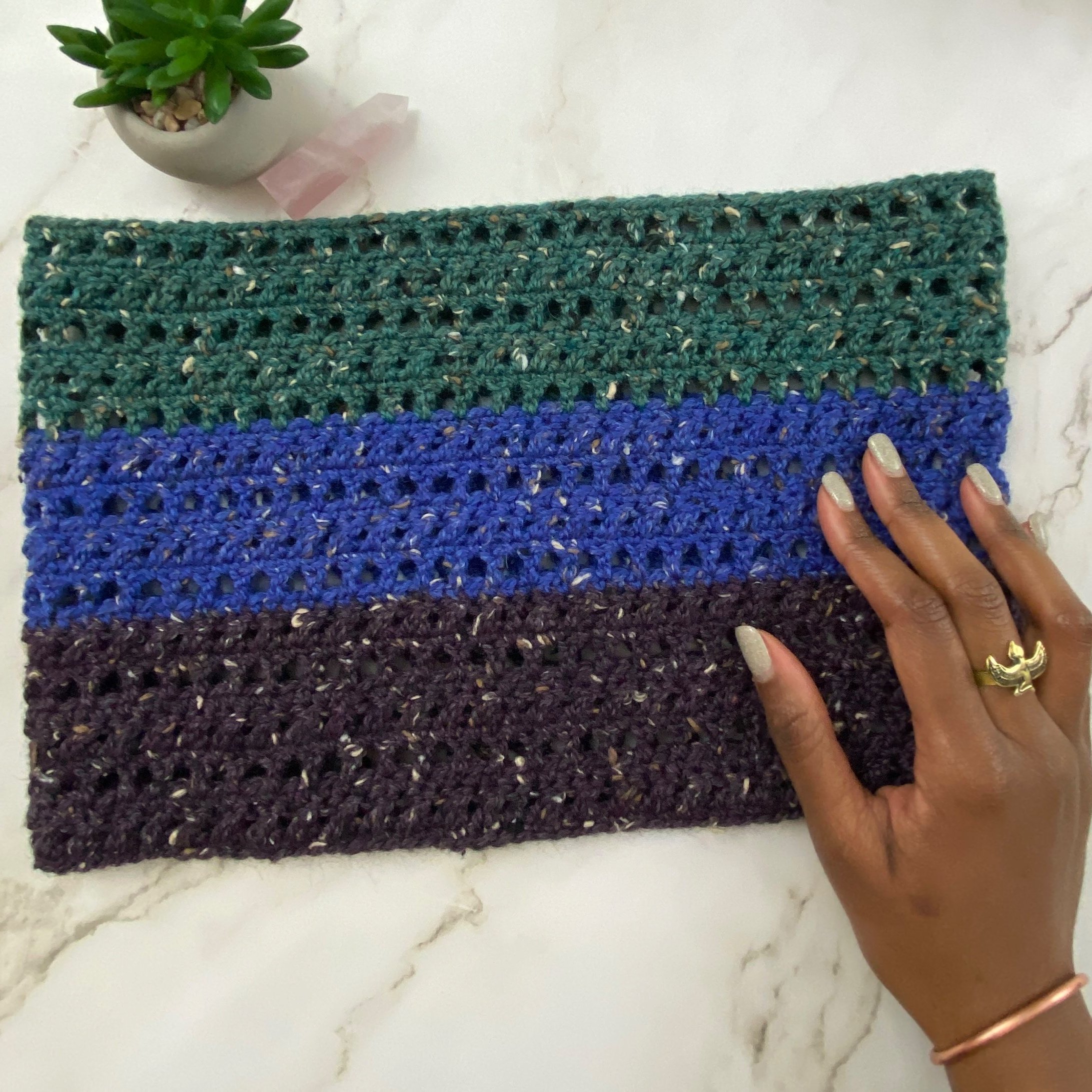 A handmade crochet cowl in shades of green, blue, black, and purple being held by a person with painted nails and wearing gold rings, on a white marble surface. A potted succulent plant and pink quartz crystal is in the corner.