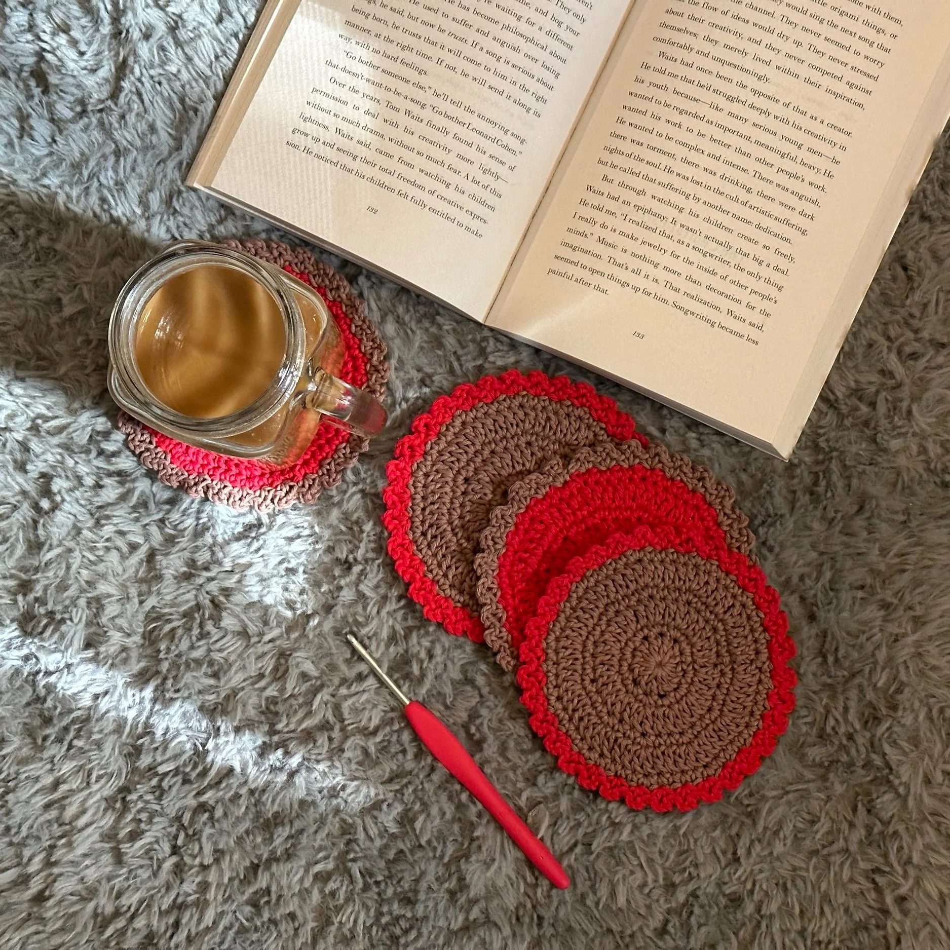A cozy scene on a gray shaggy rug featuring an open book, a crochet coaster, and a small glass mug of coffee. A red-handled crochet hook rests beside the coasters.