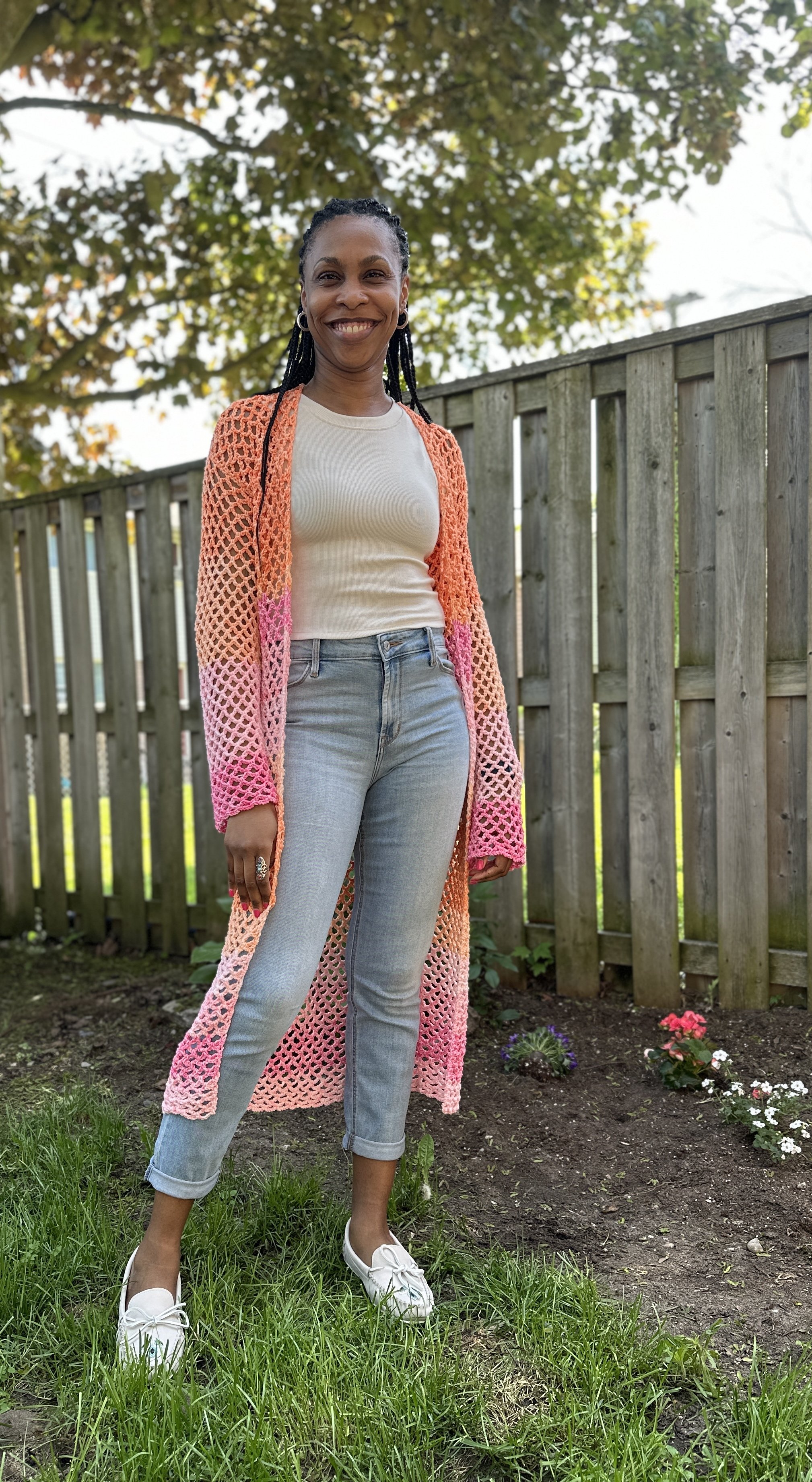 A woman standing outdoors in a garden with flowers, smiling at the camera. She is wearing a white top, light blue jeans, a pink and orange crocheted cardigan, and white sneakers. There is a wooden fence and green trees in the background.