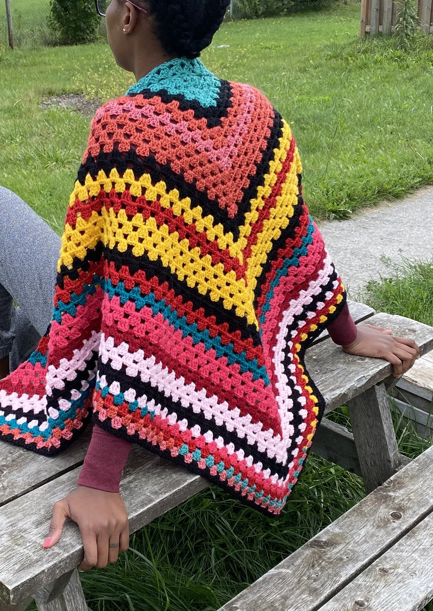 A person wearing a colorful crochet blanket with various colors and patterns, sitting on a wooden bench outdoors with green grass and a fence in the background.
