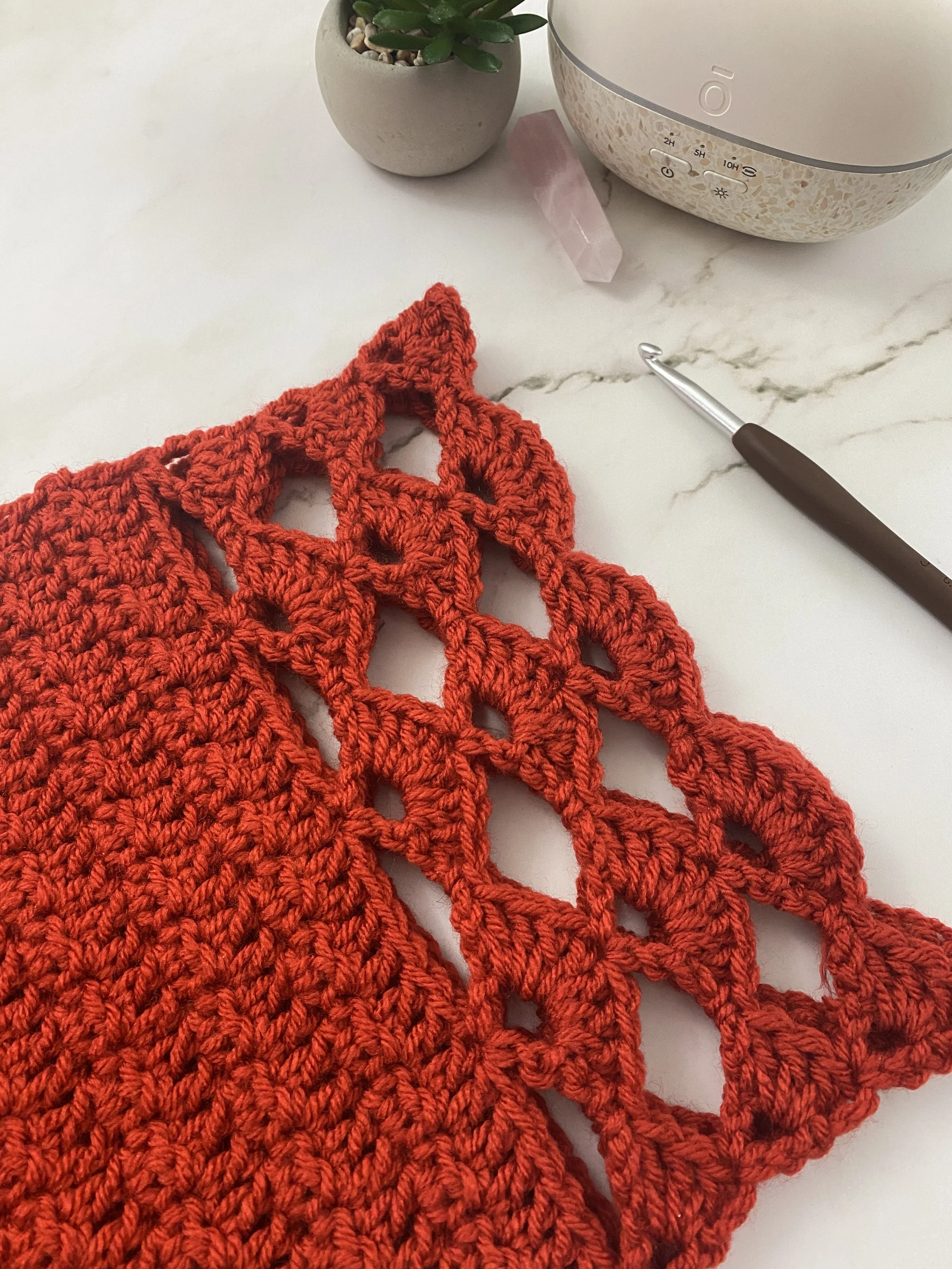 A section of orange crocheted fabric with a lace-like pattern, a small potted succulent plant, a pink quartz crystal, a white and beige ultrasonic diffuser or humidifier, and a crochet hook on a white marble surface.