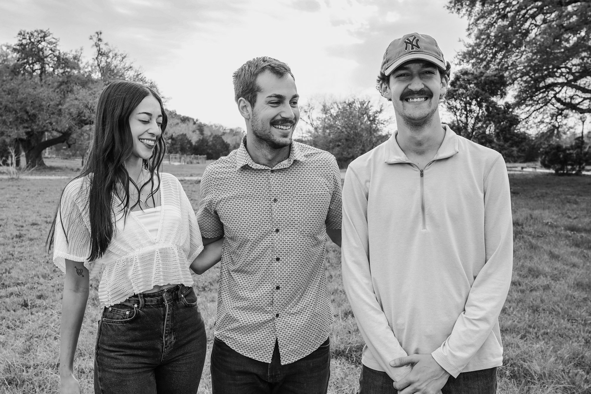 Three people, two men and one woman, standing outdoors in a park with trees in the background. They are smiling and enjoying each other's company, with the woman on the left, a man in the middle, and another man on the right wearing a cap.