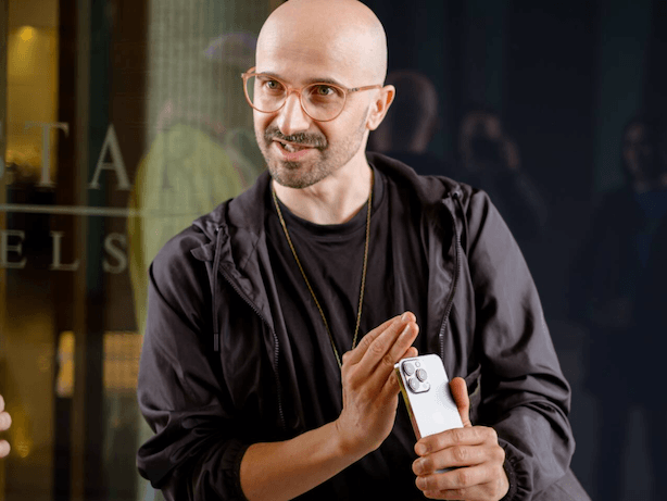A bald man with glasses holding a smartphone in front of a dark background, smiling and gesturing with his right hand.