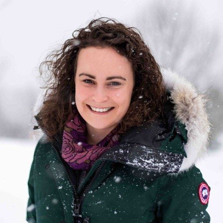 Person wearing a green parka with a fur-lined hood in a snowy outdoor setting smiling.