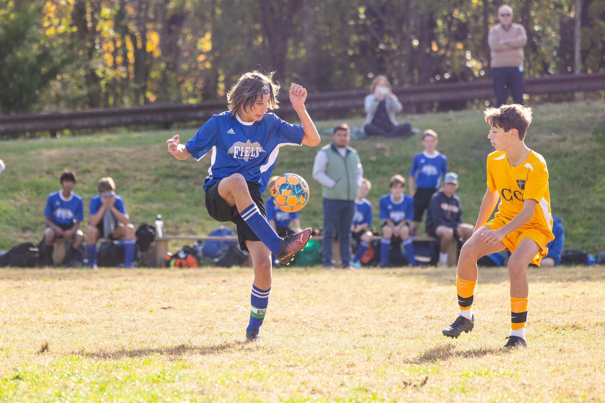 Young boys playing soccer on a grassy field during daytime, with spectators and other players sitting and standing on the sidelines, surrounded by trees with fall foliage.