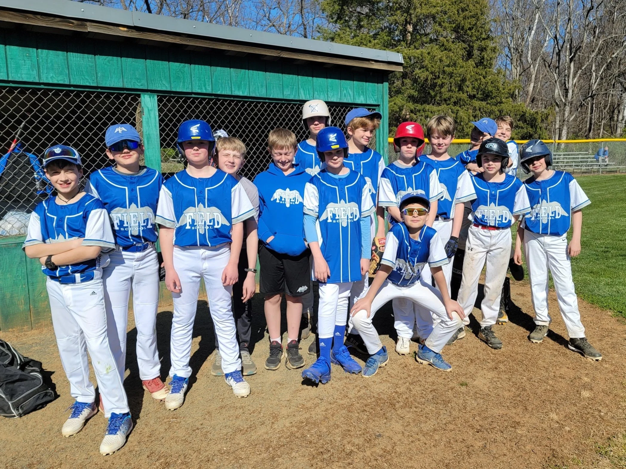 A group of young boys in blue and white baseball uniforms posing together on a baseball field. Some are wearing helmets and sunglasses, with a green dugout and trees in the background.