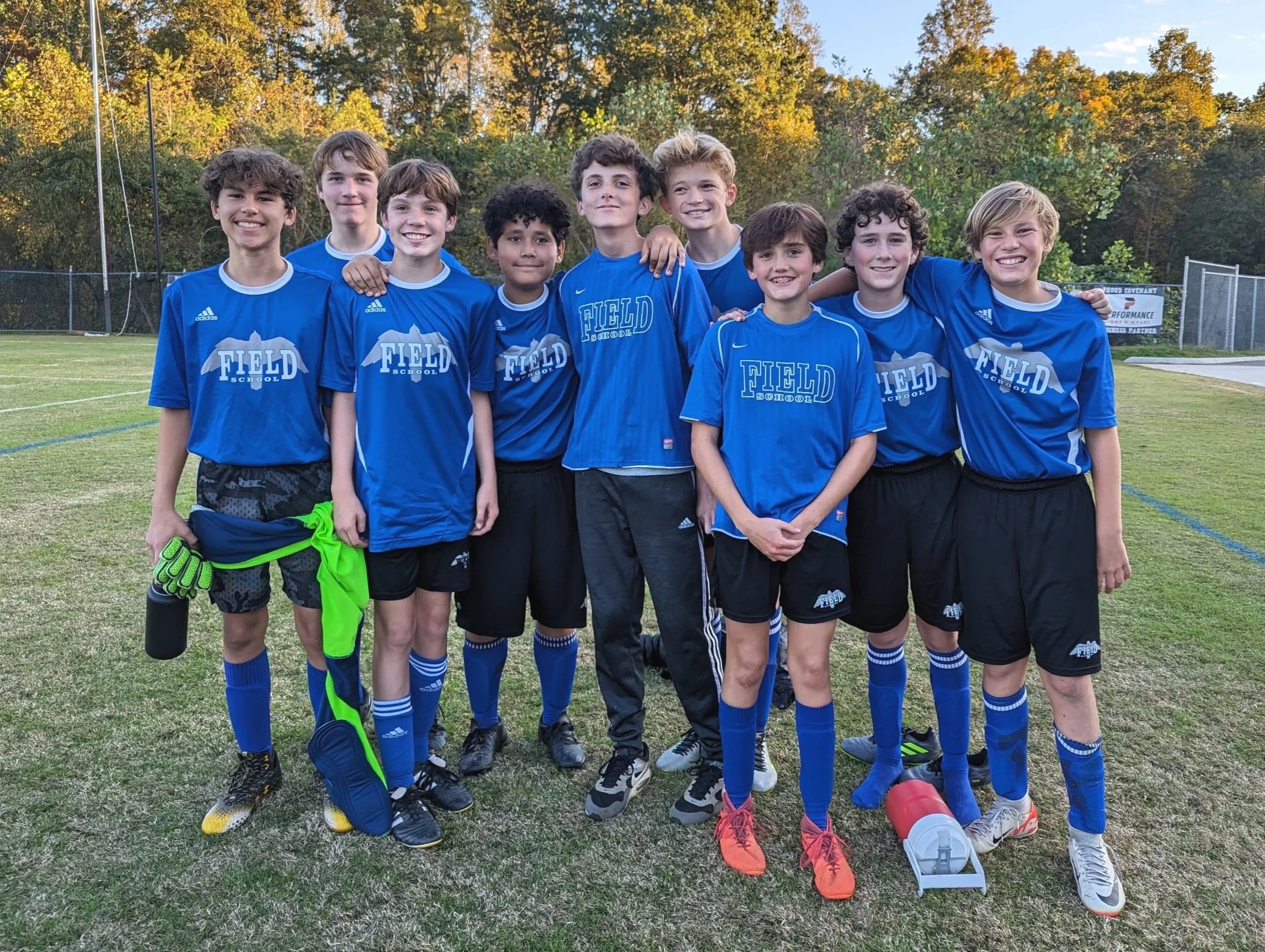 Youth soccer team wearing blue jerseys with 'Field School' logo, standing on a soccer field with trees in the background, smiling and posing for a group photo.