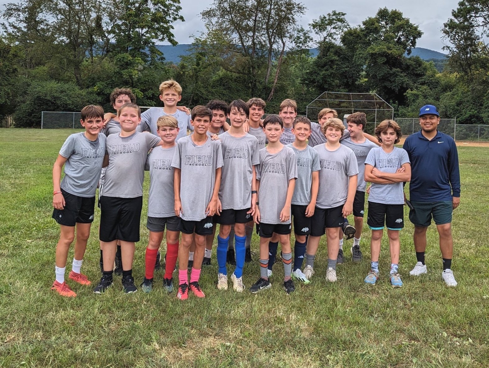 A group of young boys in gray T-shirts and shorts standing on a grassy field, posing for a photo with an adult coach, in front of a baseball/softball field with trees and mountains in the background.
