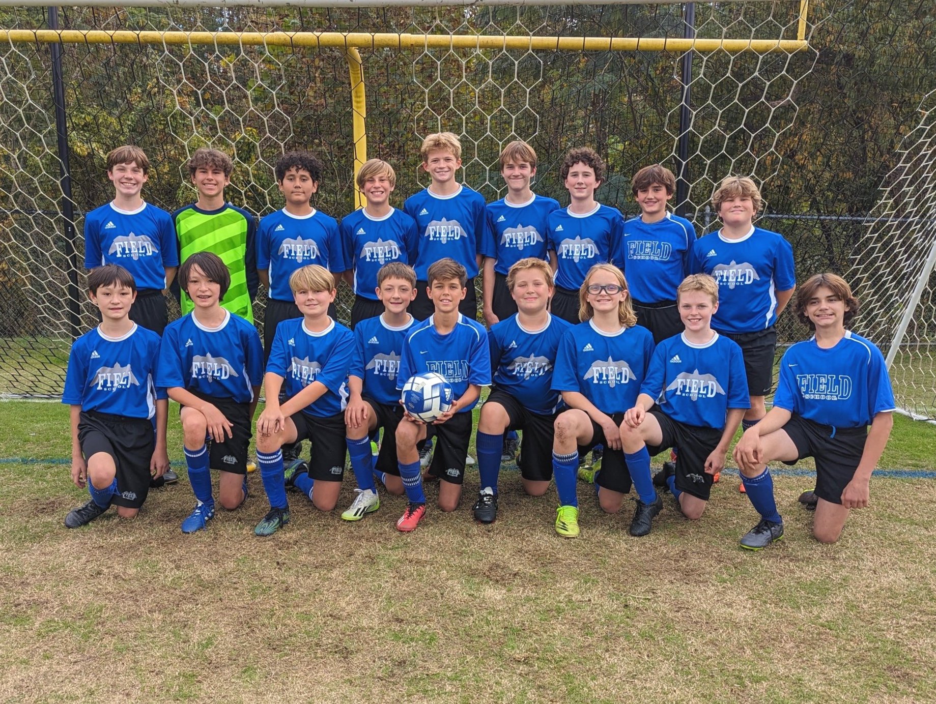 A youth soccer team wearing blue jerseys with 'FIELD' printed on them, posing for a team photo in front of a soccer goal on a grassy field, with some kids kneeling in the front row and others standing behind.
