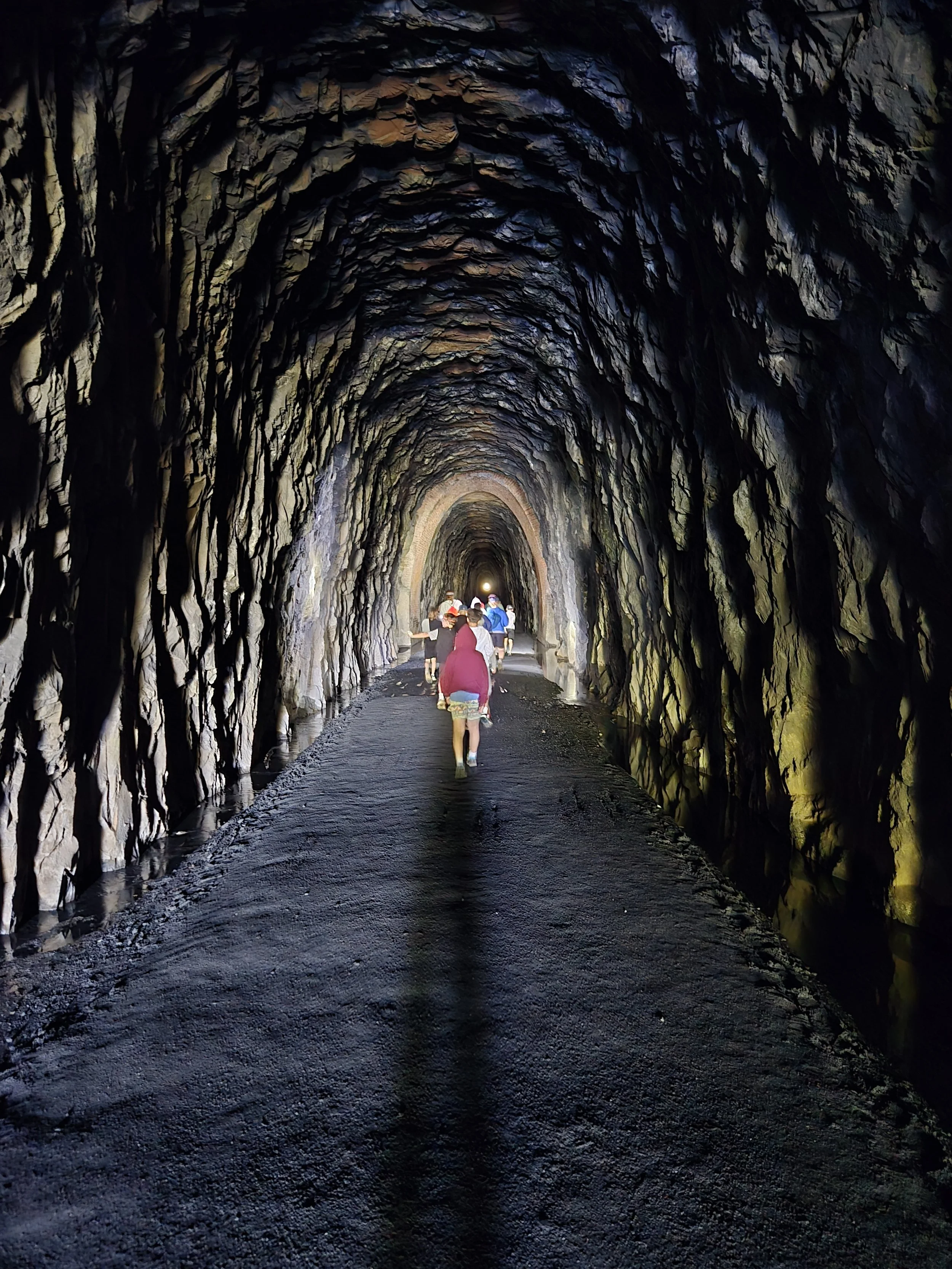 People walking inside a dark tunnel with rocky walls and a wet, gravel floor, illuminated with some lights.