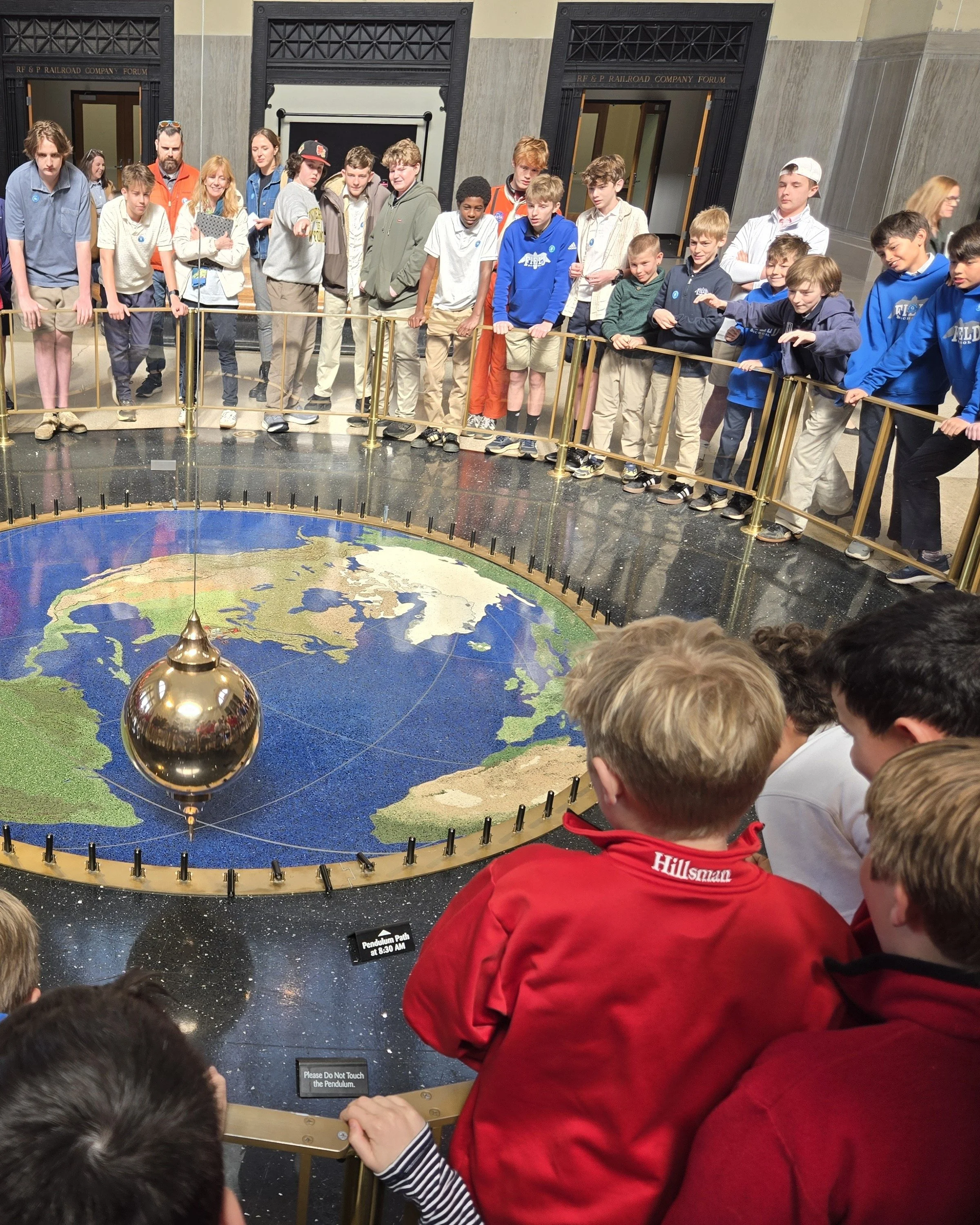 A group of children and teenagers gathered around a large, illuminated globe of the Earth. They are in an indoor museum with black and gray decor, behind a gold-colored barrier. Some children are leaning on or holding the barrier, and some are pointing or looking at the globe. There is a small sign that says 'Please Do Not Touch the Pendulum.' The scene suggests an educational or museum setting focused on geography or science.