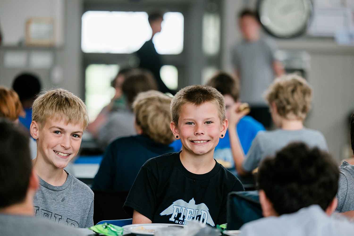 Two smiling boys with short blonde hair sitting at a cafeteria table in a school lunchroom, with other students and a teacher visible in the background.