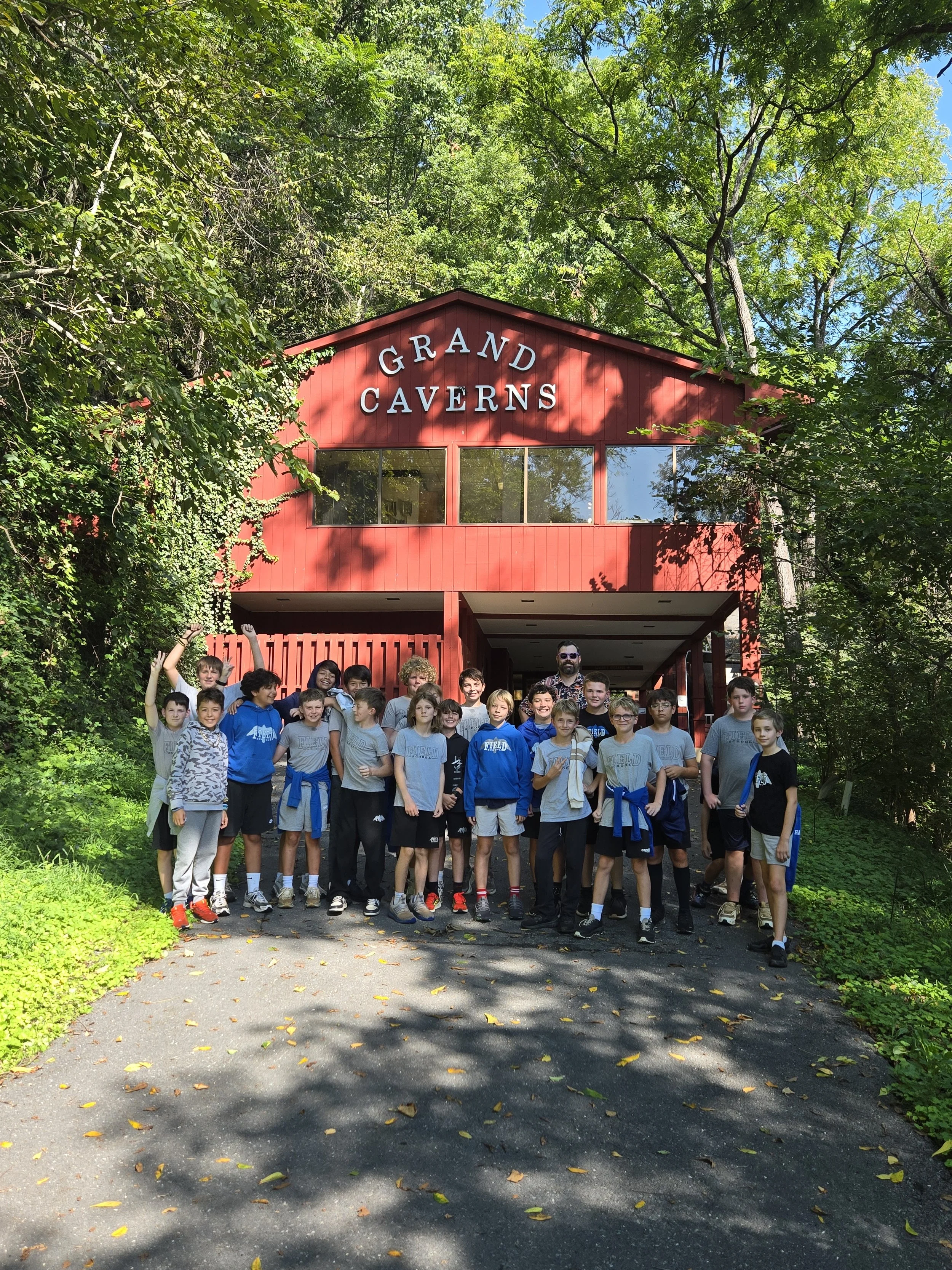 A group of children and their adult chaperone standing outside in front of a red building labeled 'Grand Caverns,' surrounded by green trees, with sunlight filtering through the leaves.