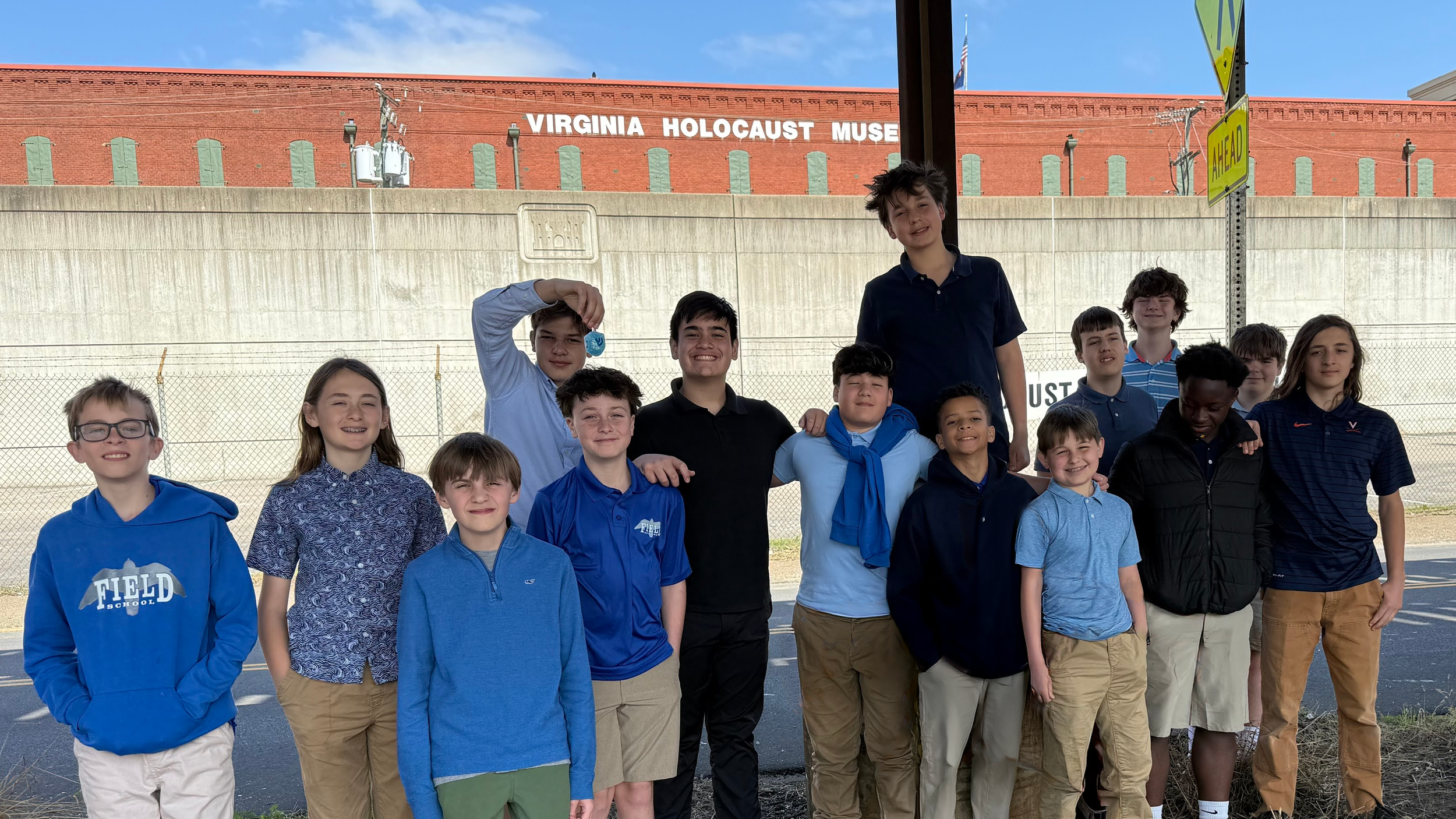 Group of young students standing outside in front of a fence with the Virginia Holocaust Museum sign on a brick building in the background.