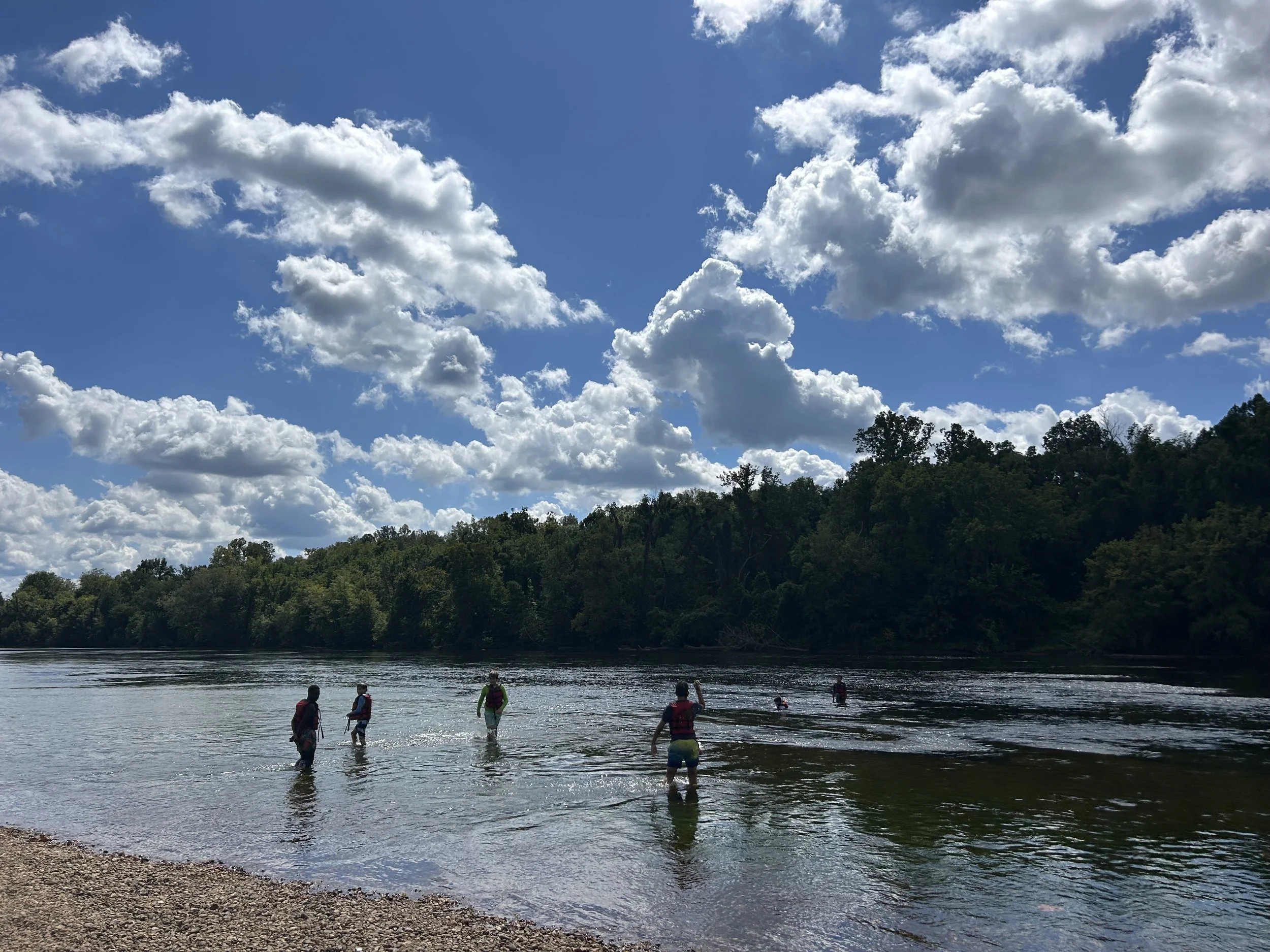 People wading in a river with a forested hillside and a partly cloudy sky in the background.
