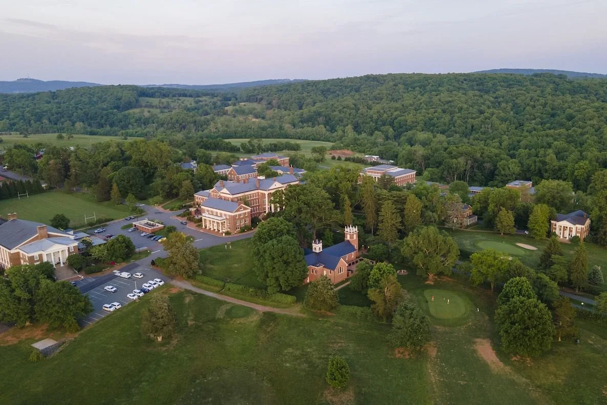 Aerial view of a college campus with large brick buildings, parking lots, surrounded by green trees and rolling hills in the background.
