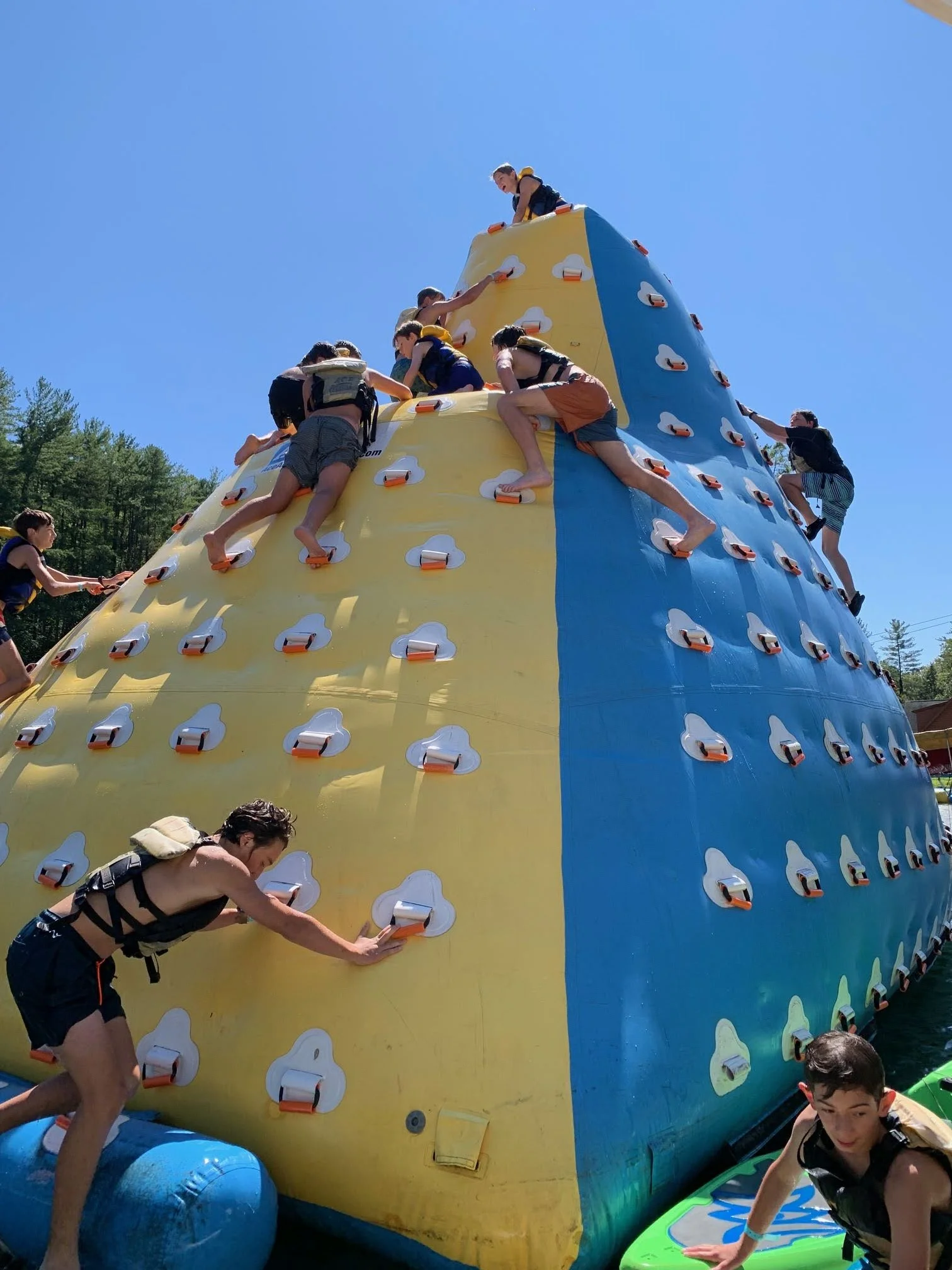 Children climbing an inflatable water obstacle course shaped like a mountain with colorful sections, set outdoors on a sunny day.