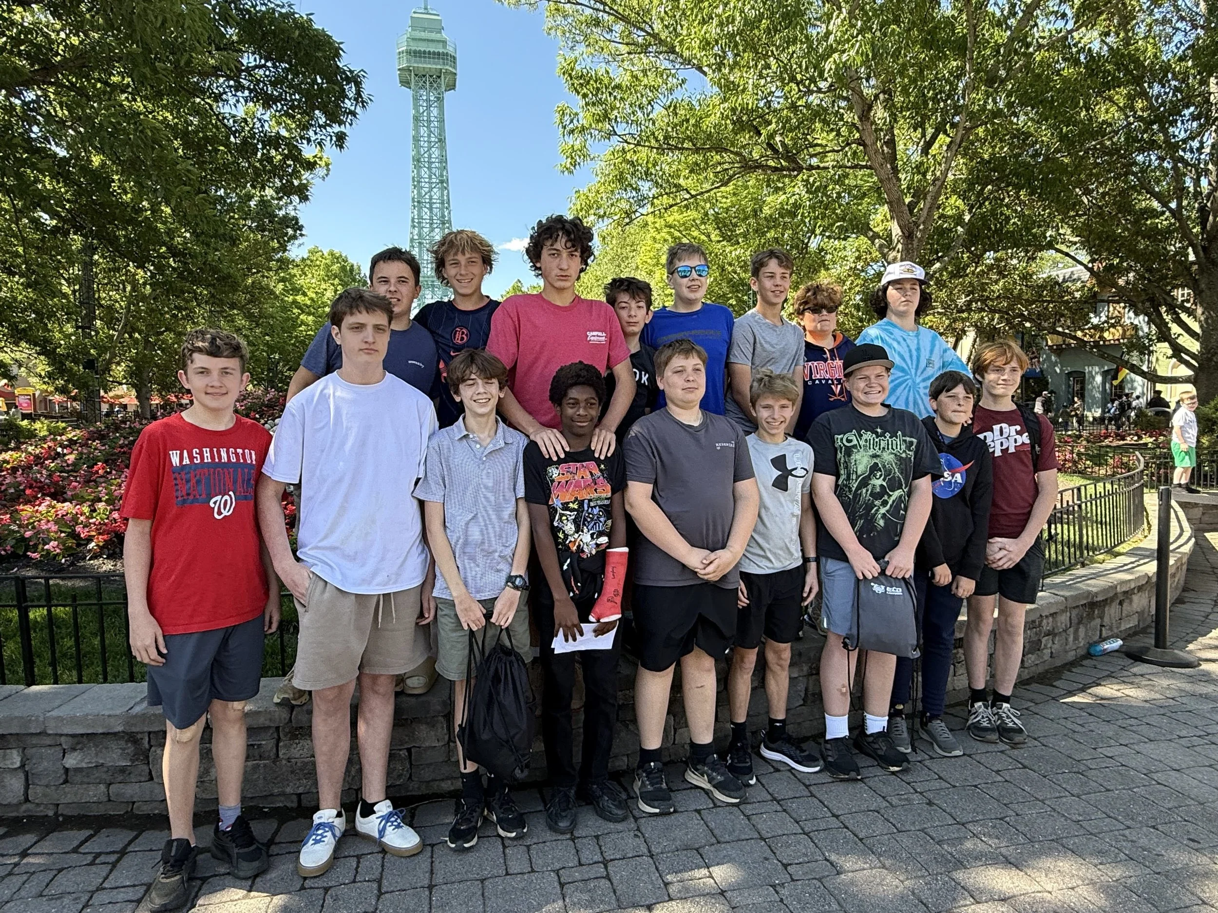 A group of boys and teenagers posing for a photo outdoors in a park. They are standing in front of a railing with trees and a tall tower in the background. It is a bright, sunny day.