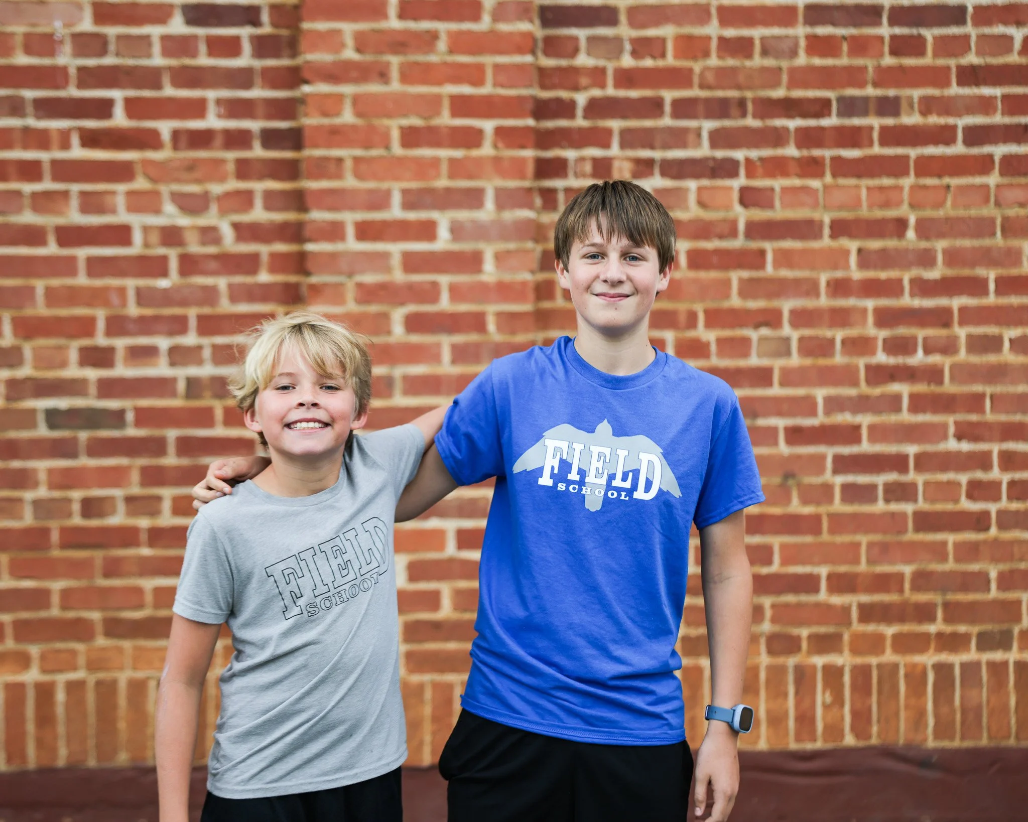 Two young boys standing in front of a brick wall, smiling, one with blonde hair wearing a gray T-shirt and the other with brown hair wearing a blue T-shirt.