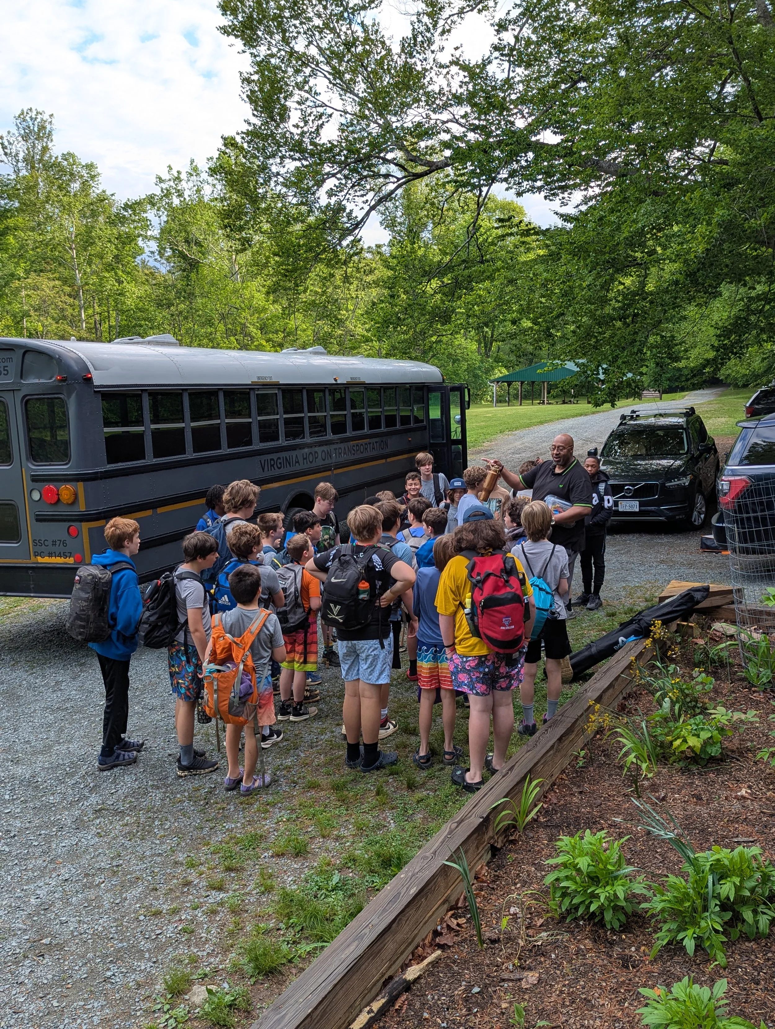 A group of children with backpacks gathered around a man who seems to be giving a demonstration outdoors in a park. There is a Virginia Hop On Transportation bus visible on the left, and a scenic background with green trees and a cloudy sky.