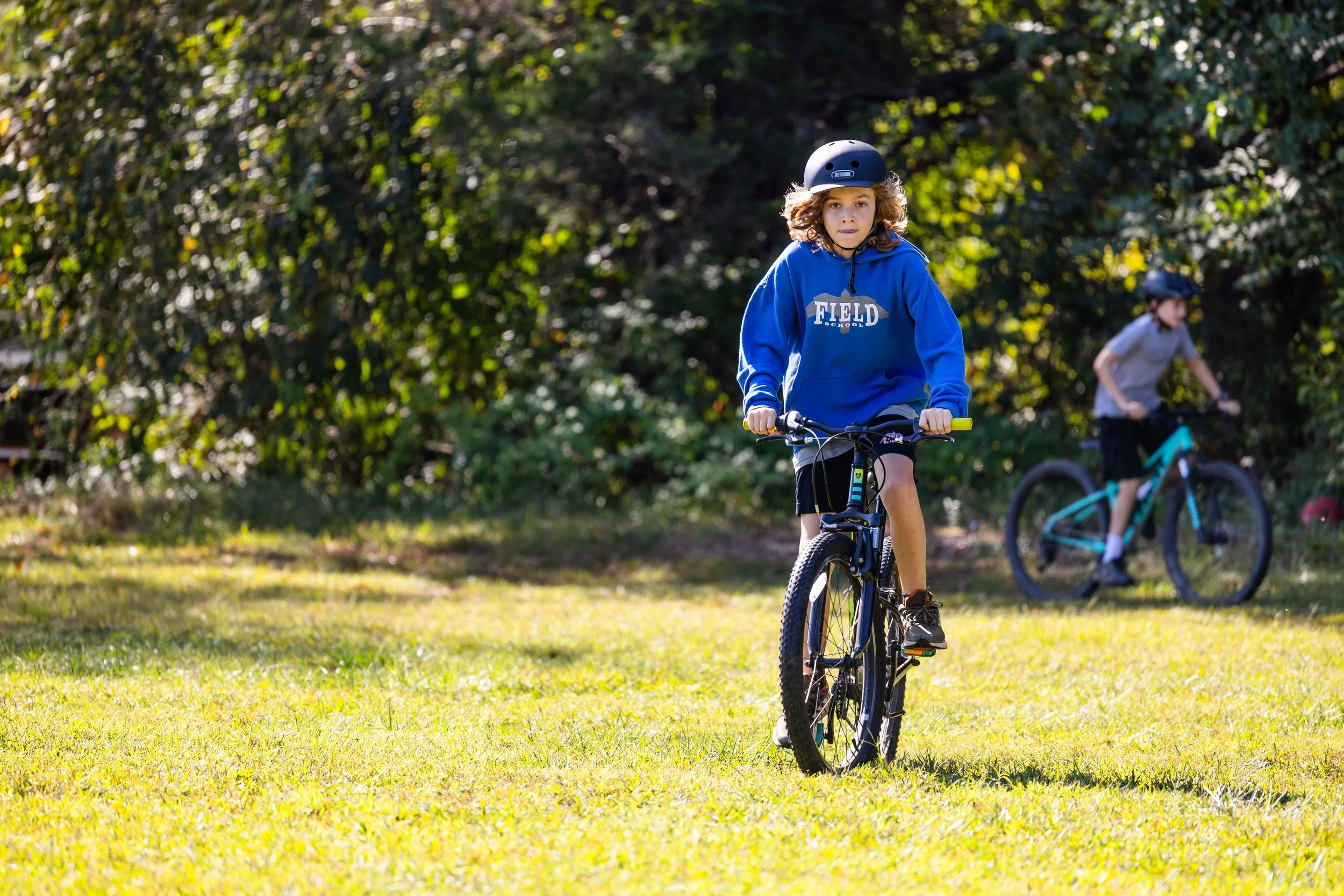 Young boy wearing a blue hoodie and helmet riding a mountain bike on grass, with another boy in the background also on a bike, near trees.