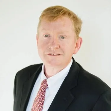 A man with red hair and fair skin wearing a black suit, white shirt, and red patterned tie against a white background.