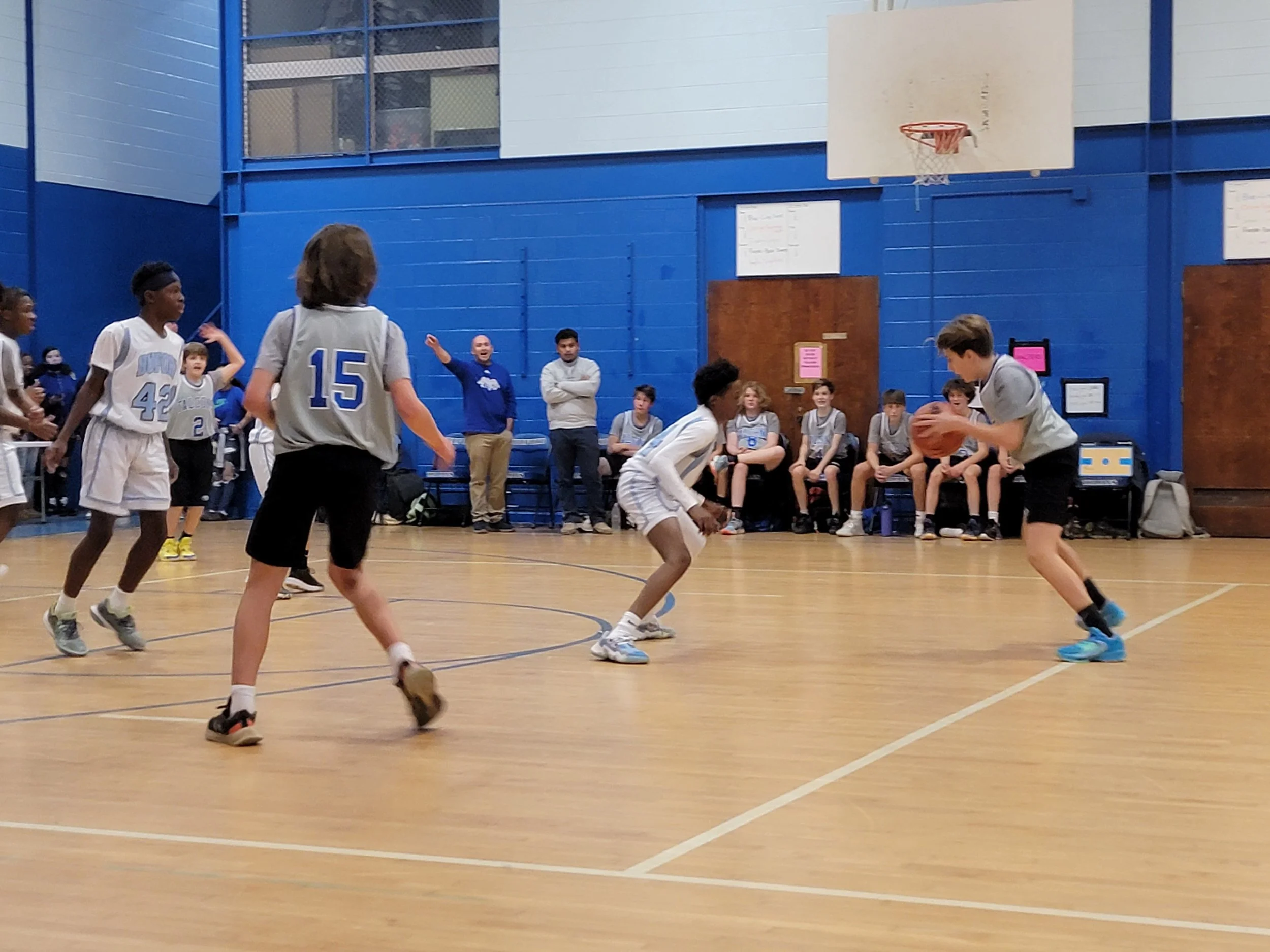 Young girls playing basketball inside a gym, with some players sitting on the bench and coaches standing in the background.