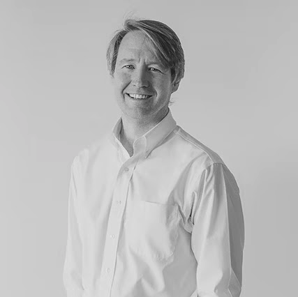 Black and white portrait of a smiling man with straight hair, wearing a button-up shirt.