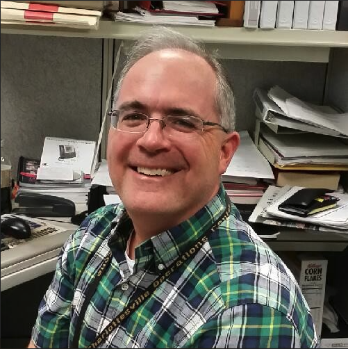 A smiling man wearing glasses and a plaid shirt in an office setting with shelves of papers and office supplies behind him.
