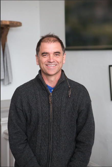 A smiling man in a dark gray zip-up sweater standing indoors with a white wall and framed picture in the background.