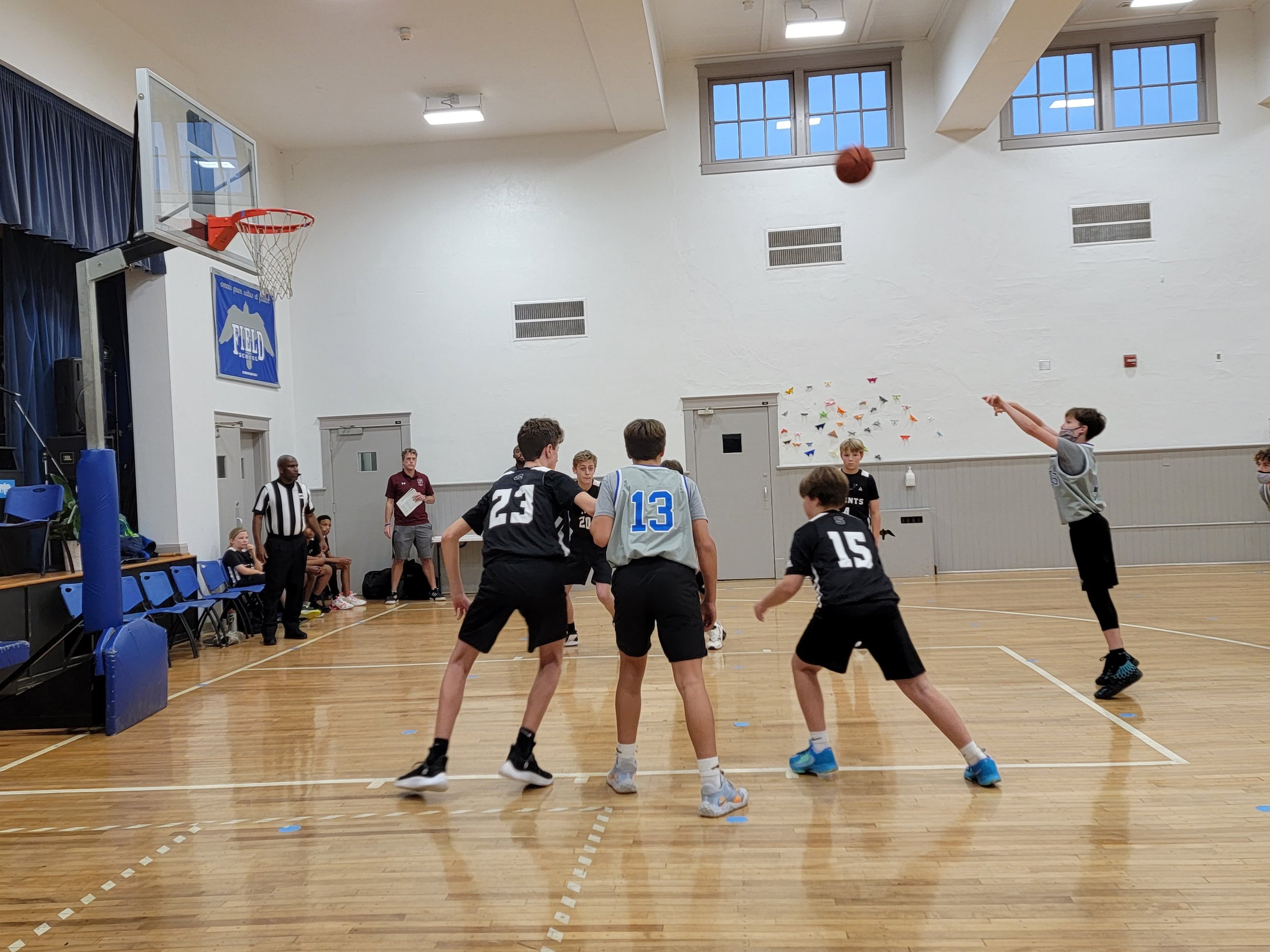 A group of young boys playing basketball indoors, with some attempting to shoot and others defending, while a referee and spectators watch from the sidelines.