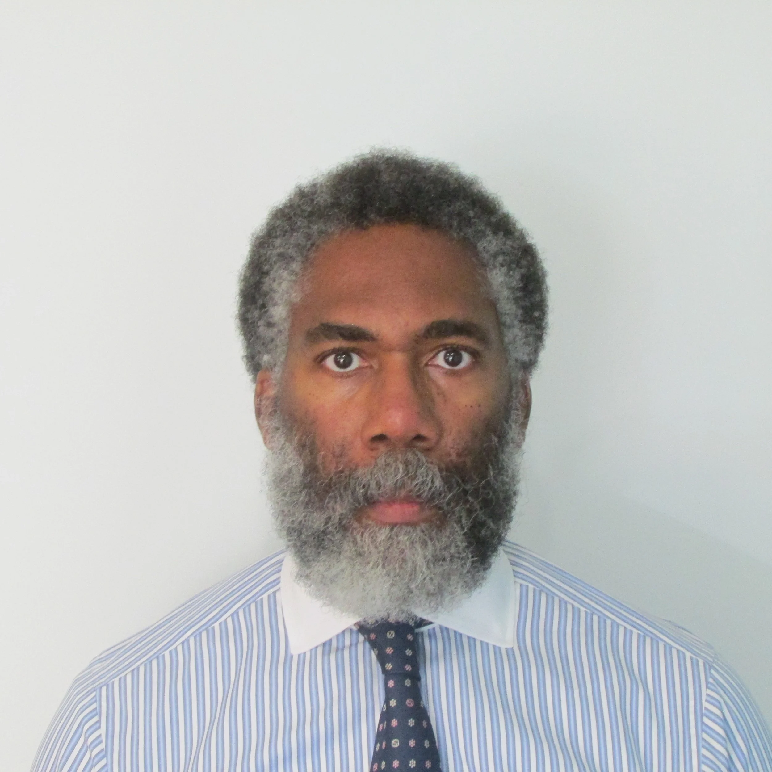 Portrait of a middle-aged man with gray hair and beard, wearing a striped shirt and dark patterned tie, standing against a plain light-colored background.