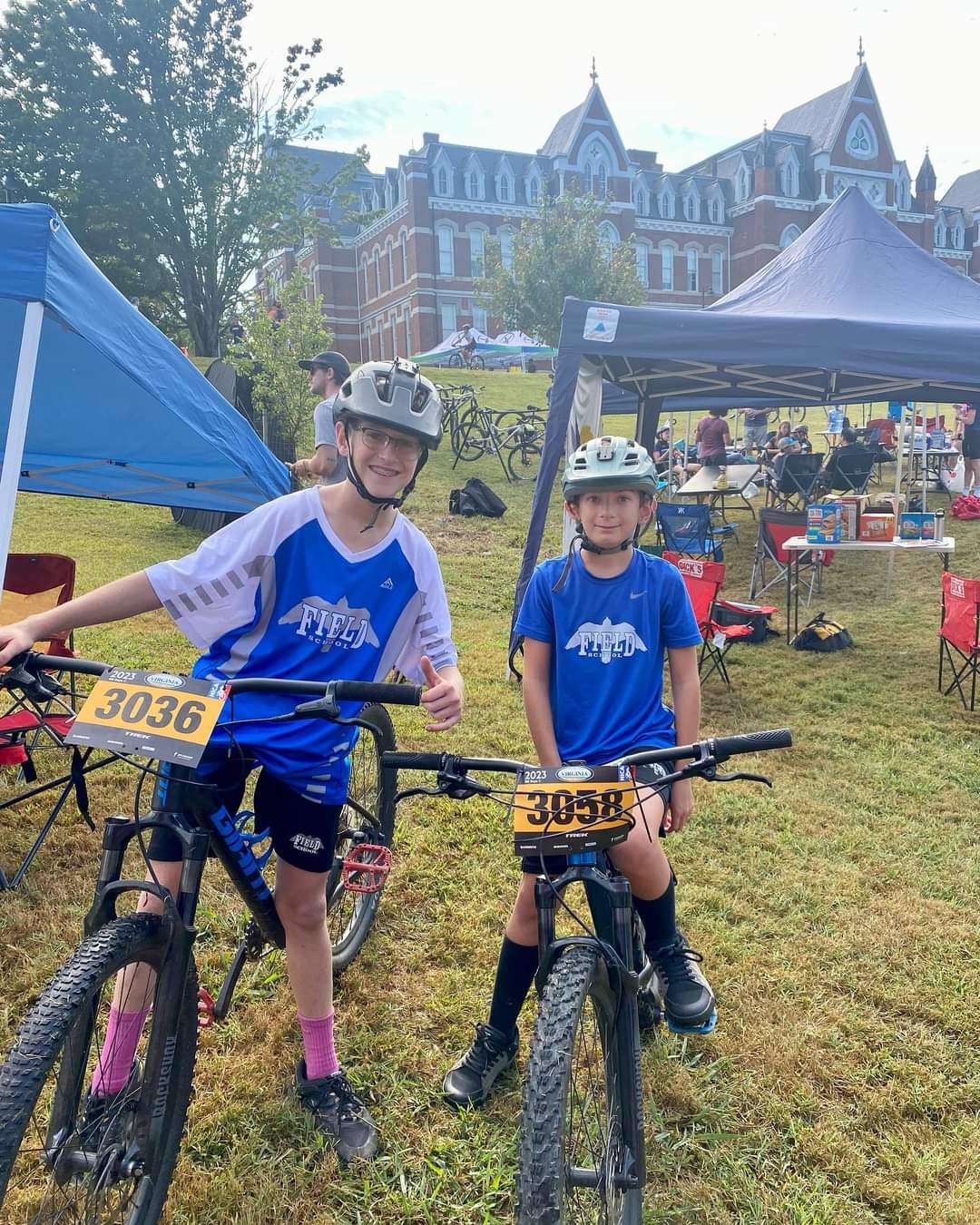 Two young cyclists wearing helmets and blue sports jerseys standing with their bikes at an outdoor event, with a historic building in the background, tents, and other people in the distance.