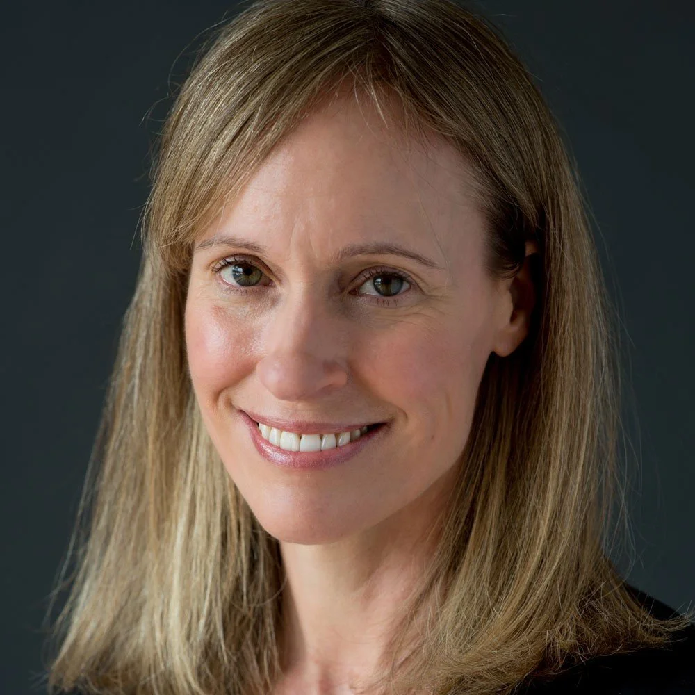 Close-up portrait of a woman with long, light brown hair, smiling slightly, wearing a dark top against a dark background.