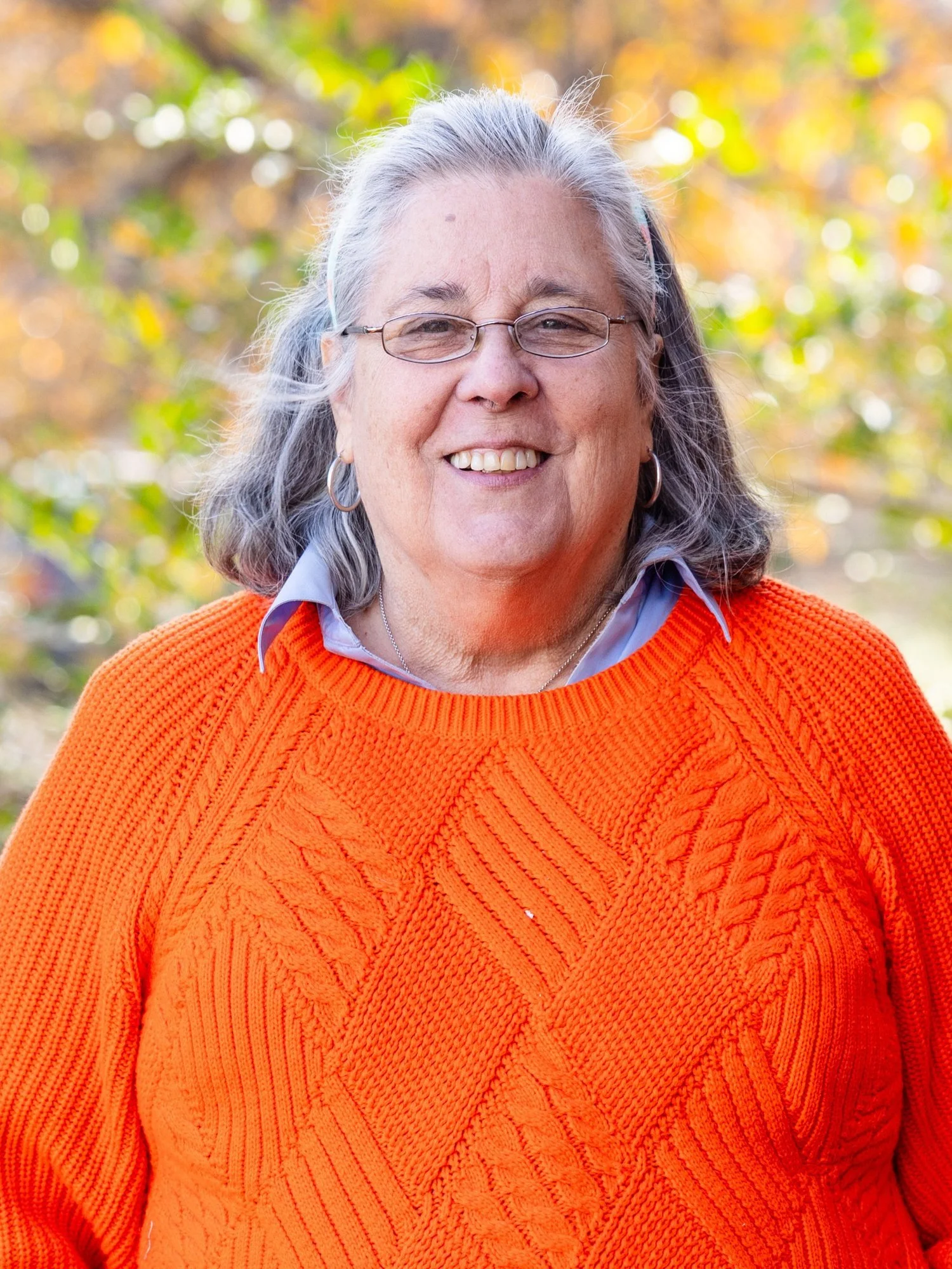 A smiling woman with glasses and hoop earrings outdoors in autumn, wearing an orange sweater over a gray collared shirt.