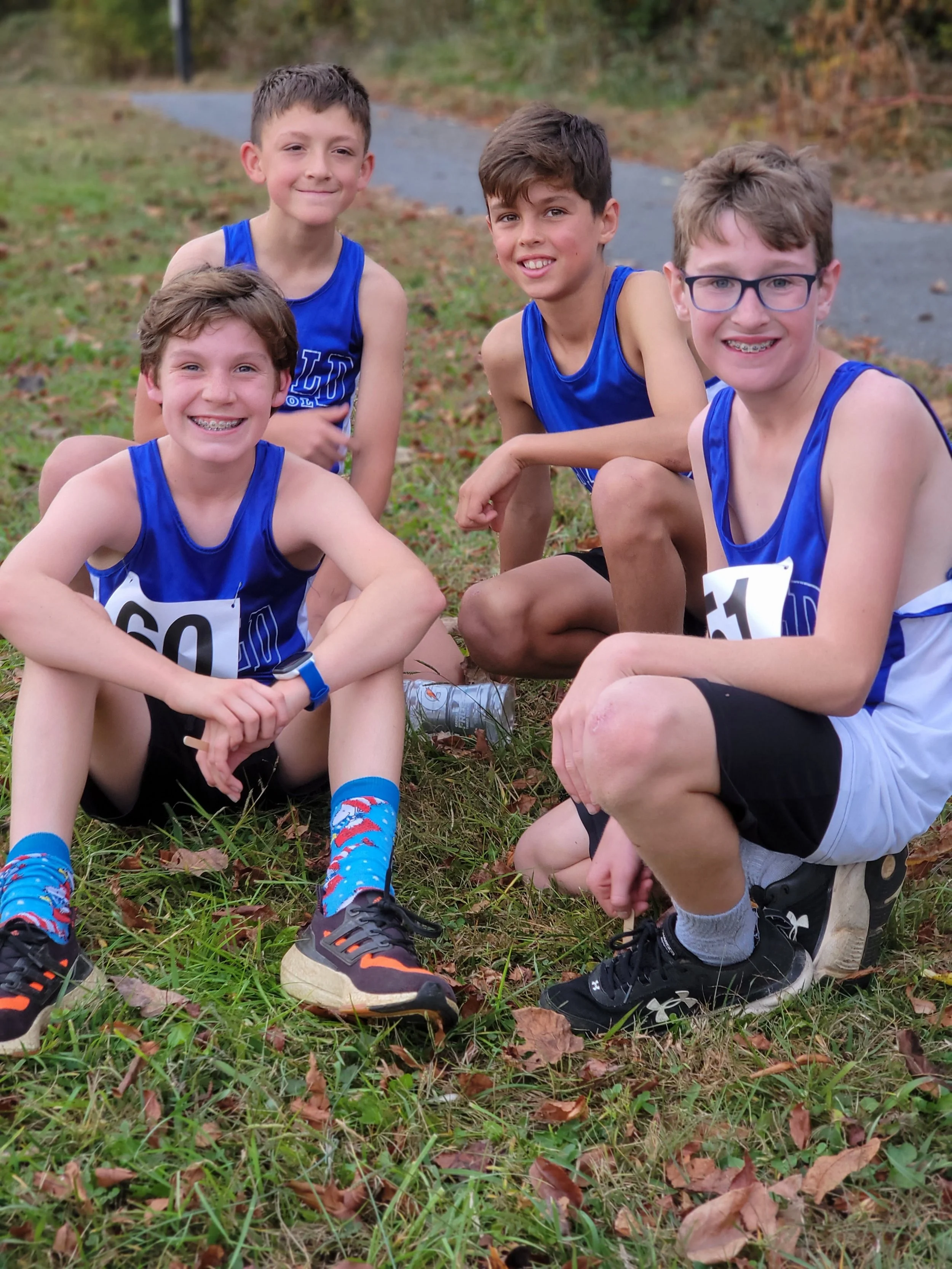 Group of five young boys in blue running uniforms squatting outside on grass with fallen leaves, smiling at the camera.