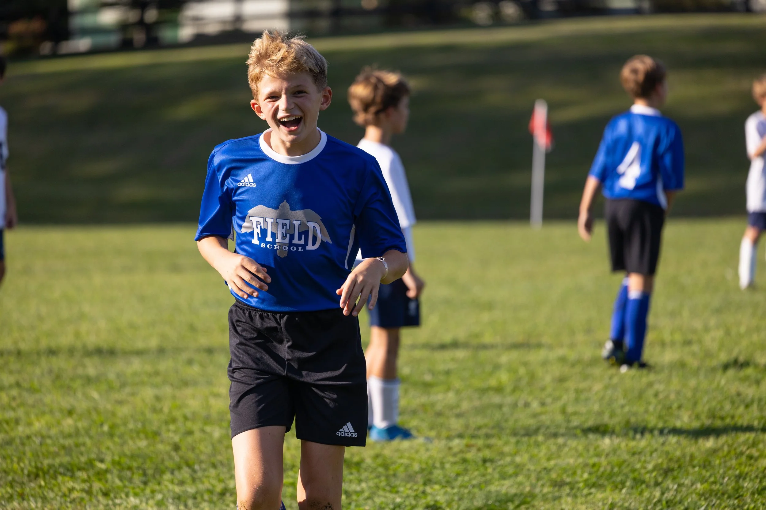 A young boy in a blue soccer jersey smiling and running on a grassy field, with other children in soccer uniforms in the background.
