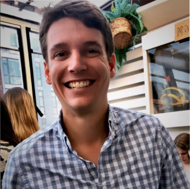 A young man smiling at a restaurant or cafe with large windows and a decorative plant in the background.